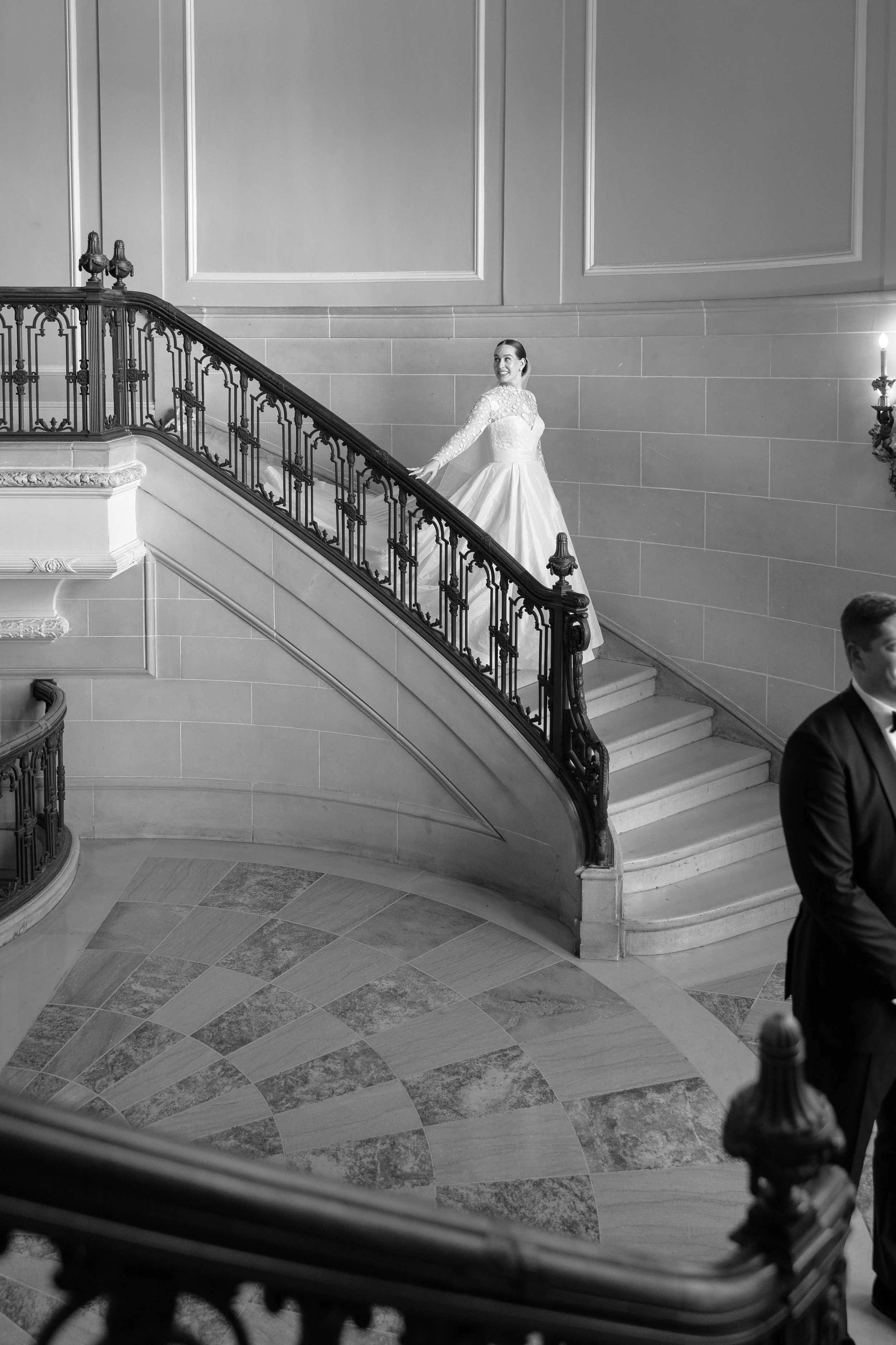 A woman in a wedding dress standing on a staircase, smiling and looking to her right, with a man in a suit partially visible in the foreground.