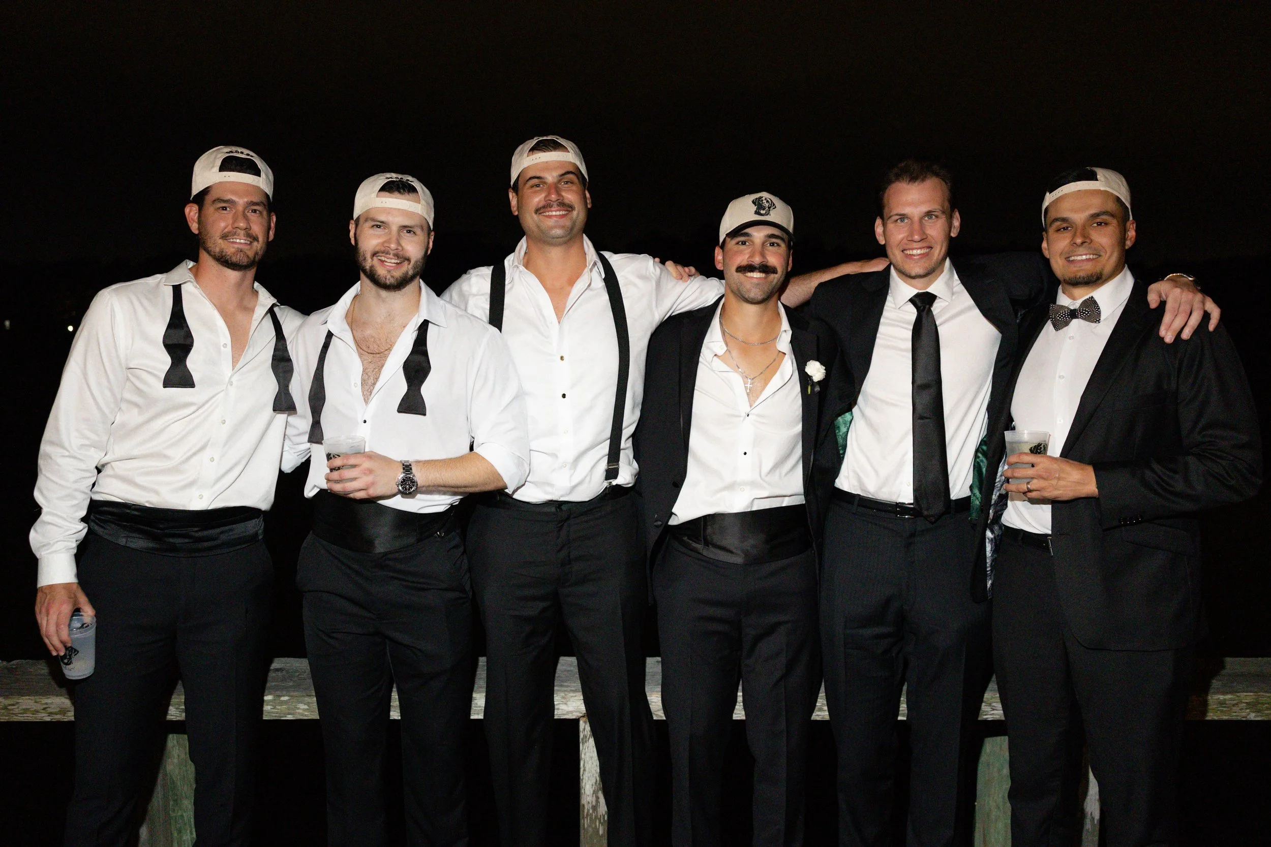 Group of seven men dressed in formal attire, smiling and standing closely together outdoors at night, some with their arms around each other.