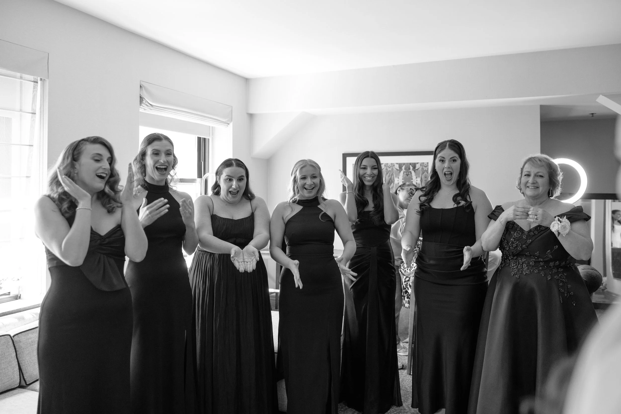 Group of women in formal dresses celebrating indoors, smiling and reacting excitedly.