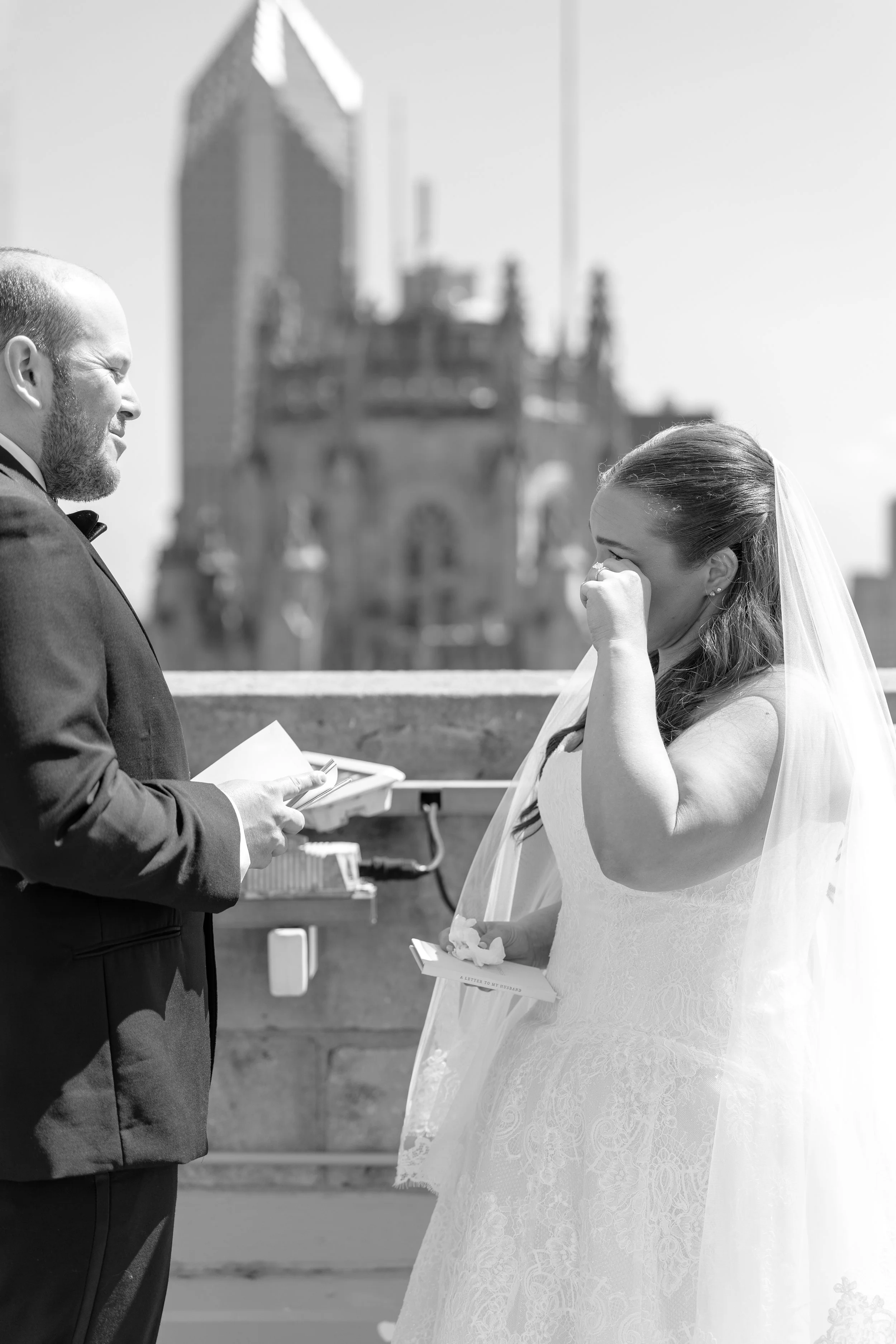 A bride and groom standing face to face outdoors, with the groom holding a note paper and the bride wiping tears from her eyes.