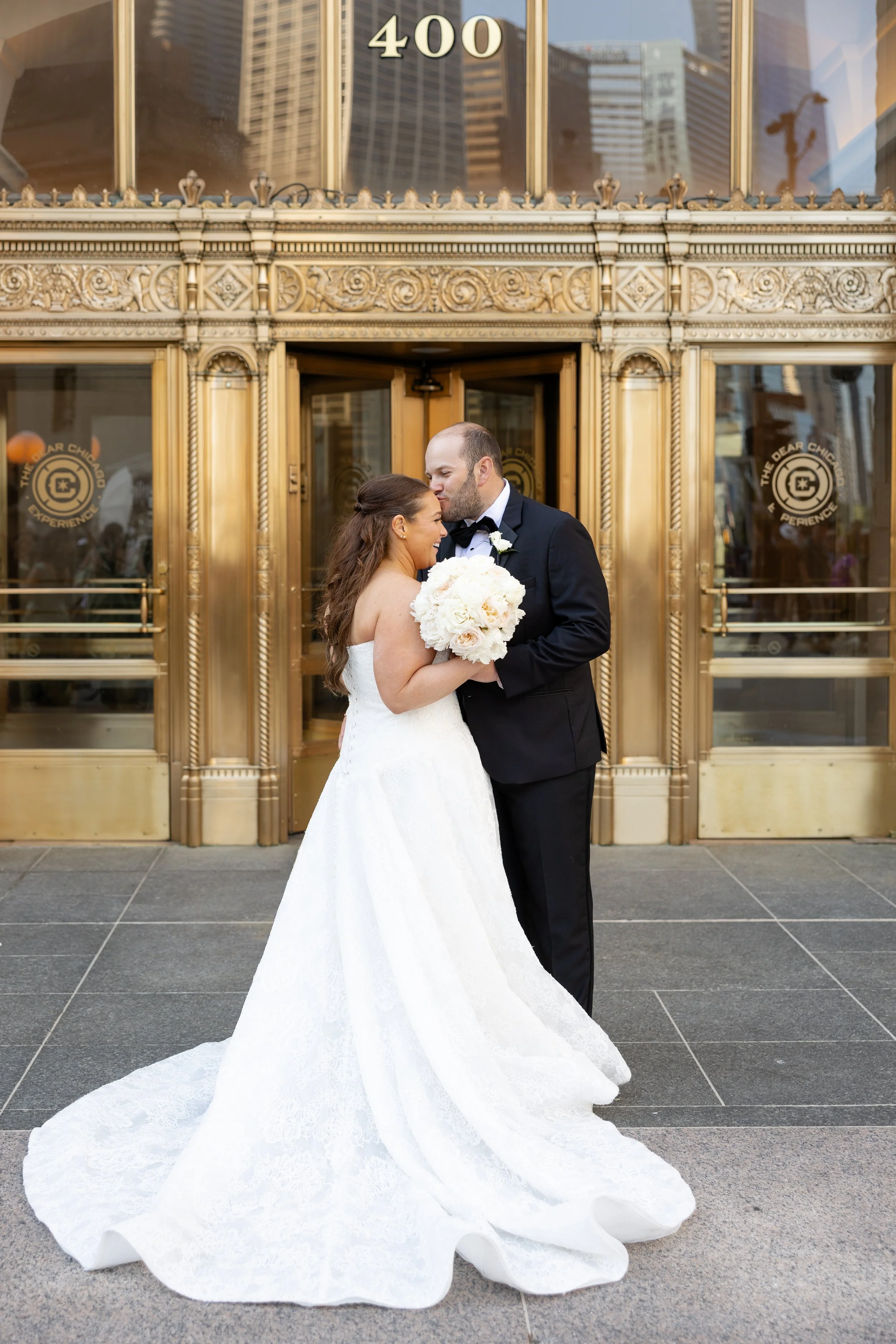 A bride and groom share a kiss outside a building with ornate gold doors. The bride is wearing a white wedding gown with a long, flowing train and holding a bouquet of white flowers. The groom is in a black tuxedo with a bow tie and white shirt. The 