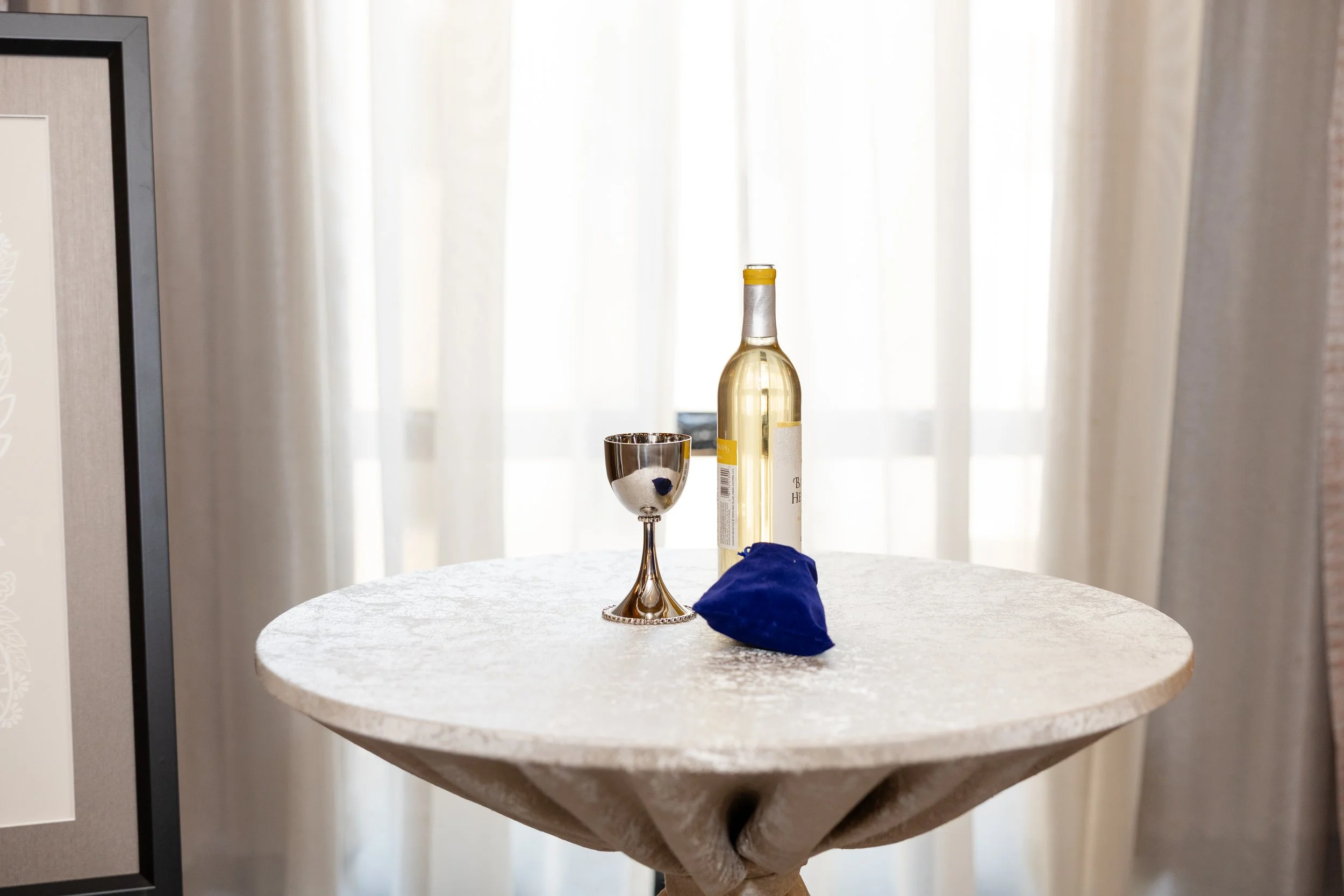 Bottle of white wine, silver goblet, and dark blue cloth on a round table with a textured white tablecloth, in front of sheer curtains.