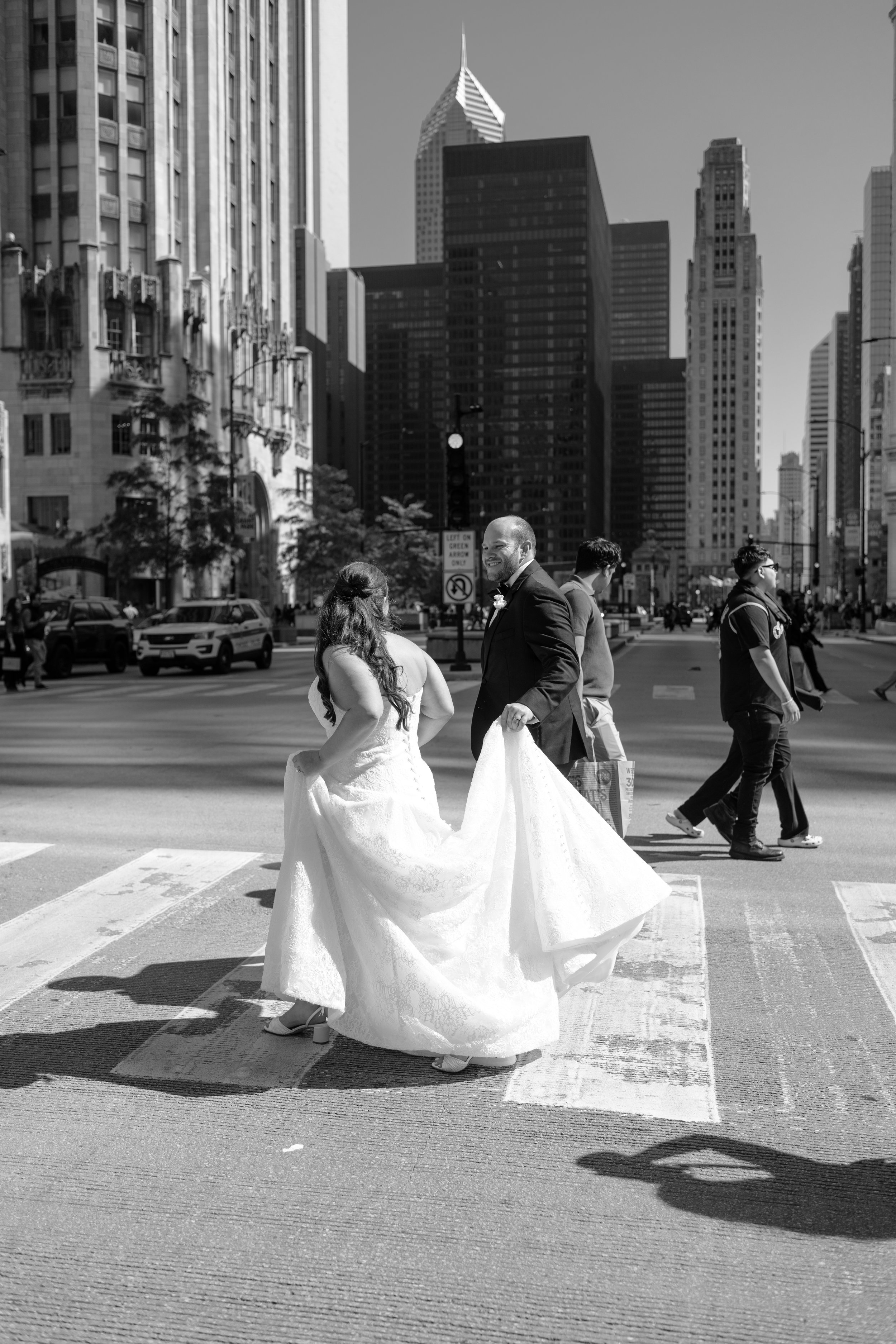 Black and white photo of a bride and groom holding hands in a city crosswalk with tall buildings in the background.