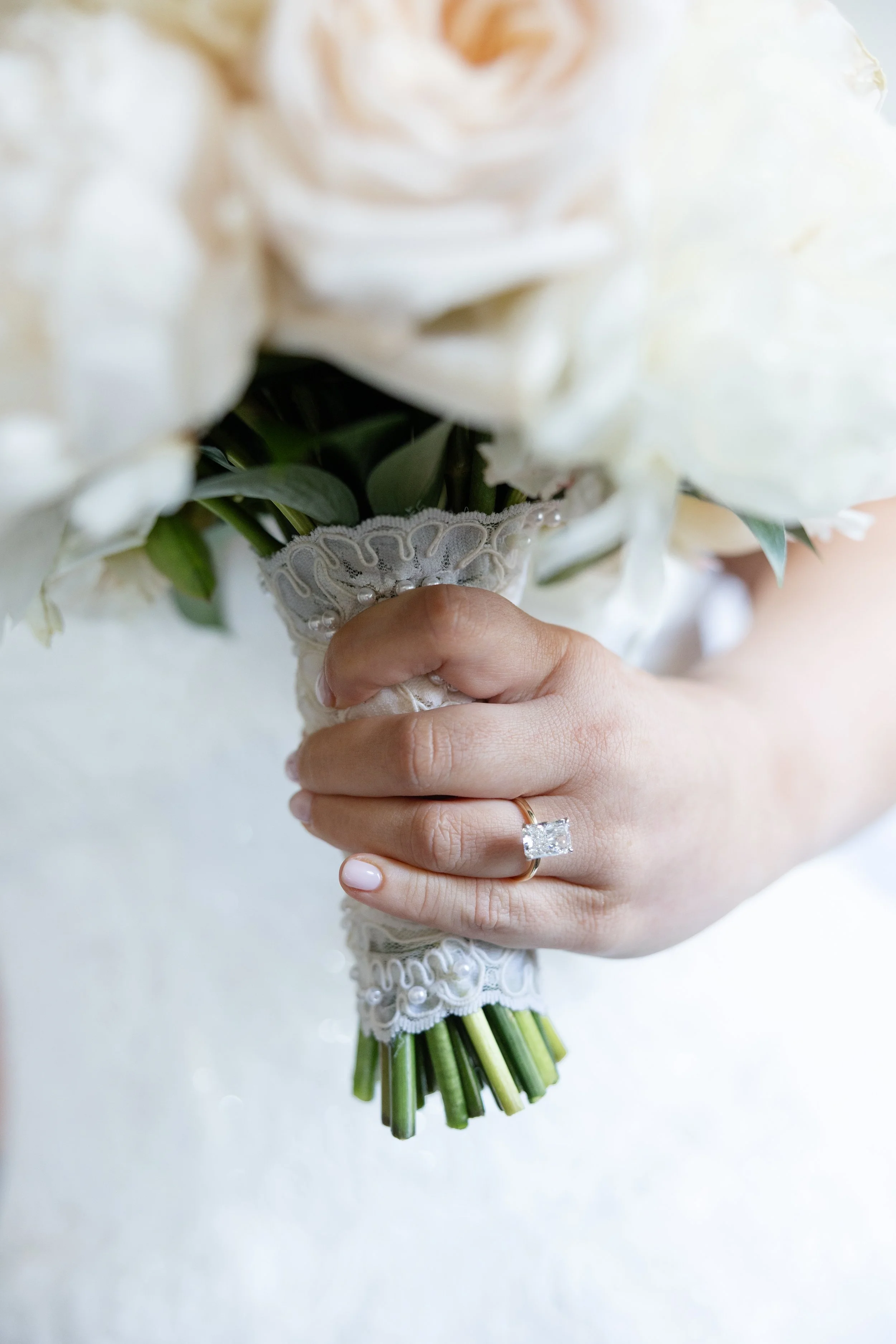 Close-up of a hand holding a bridal bouquet of white peonies with green stems and leaves, wrapped with lace and pearl accents, with a large engagement ring on the ring finger.