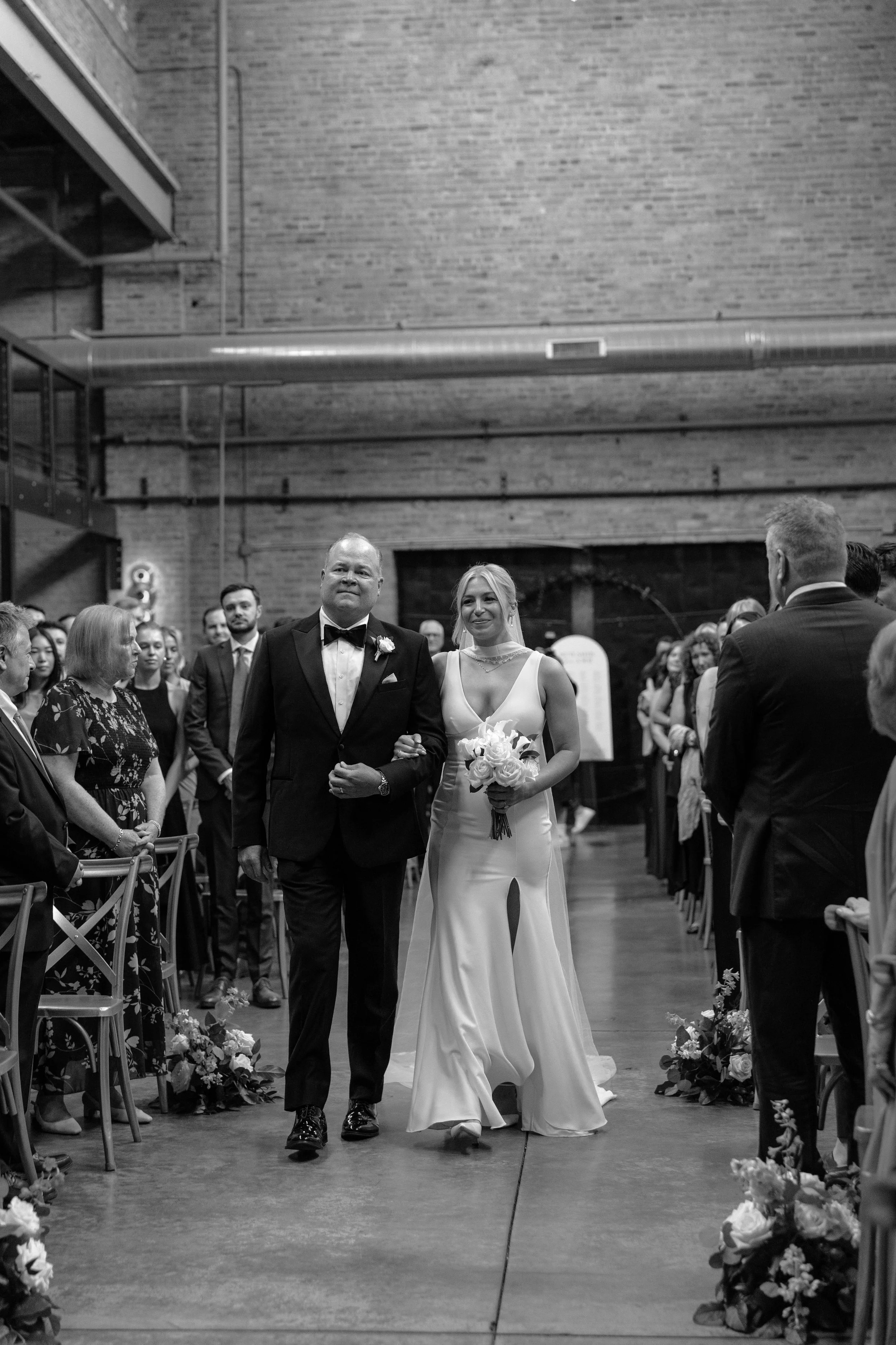 A bride and a groom walking down the aisle at their wedding ceremony, surrounded by guests in a brick-walled venue.