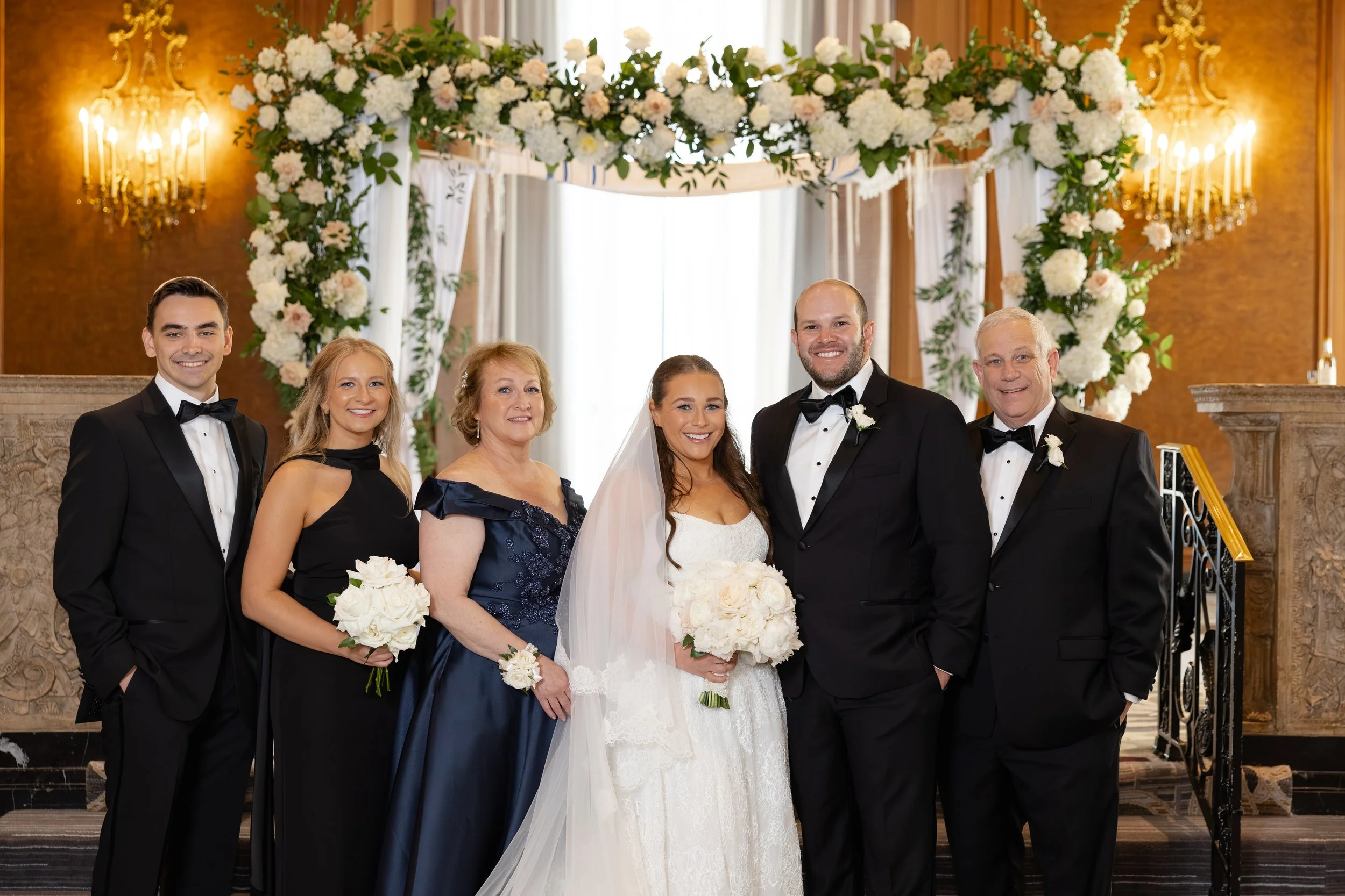 A wedding group photo featuring a bride, groom, and four other well-dressed people standing in front of a floral wedding arch. The background includes ornate wall sconces and elegant drapery.