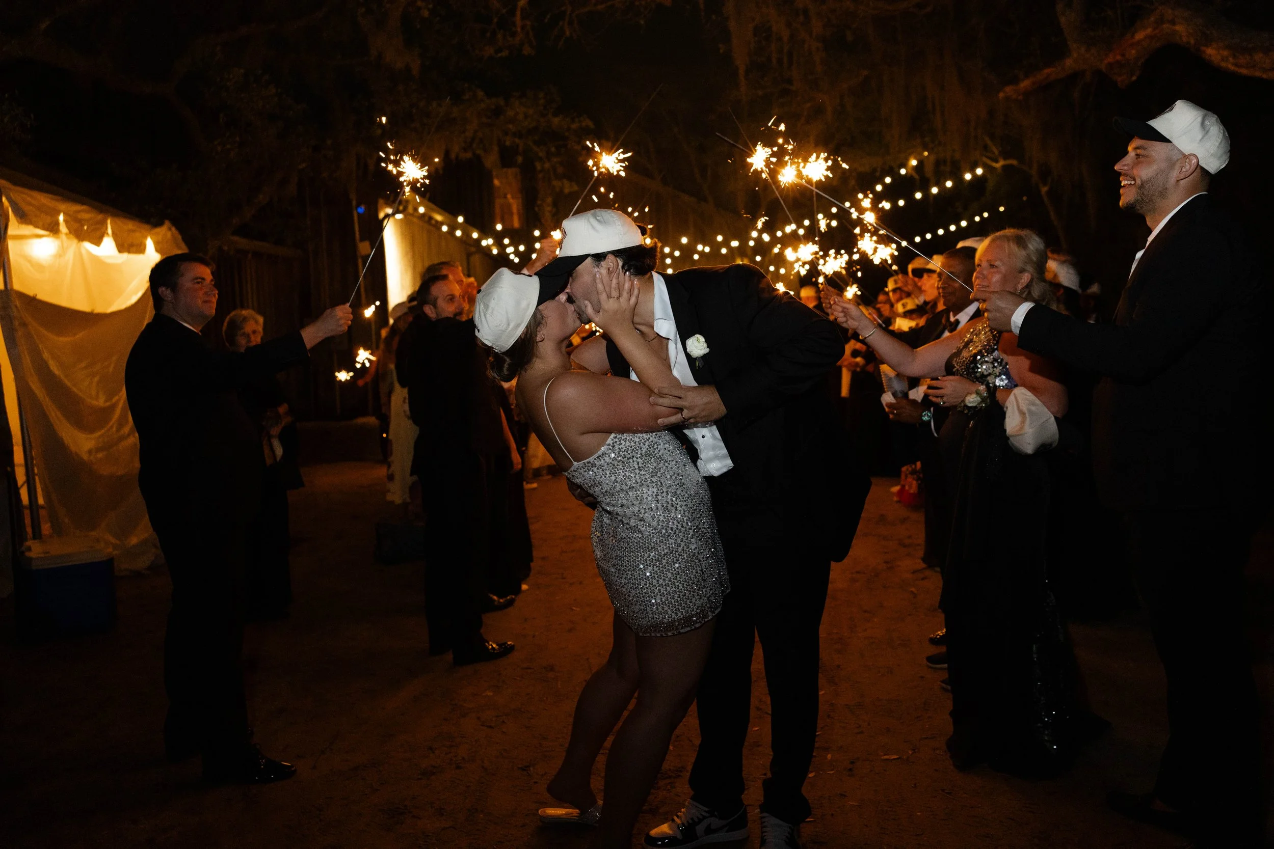 A couple kisses at night during a celebration, possibly a wedding, while surrounded by guests holding sparklers and string lights overhead.