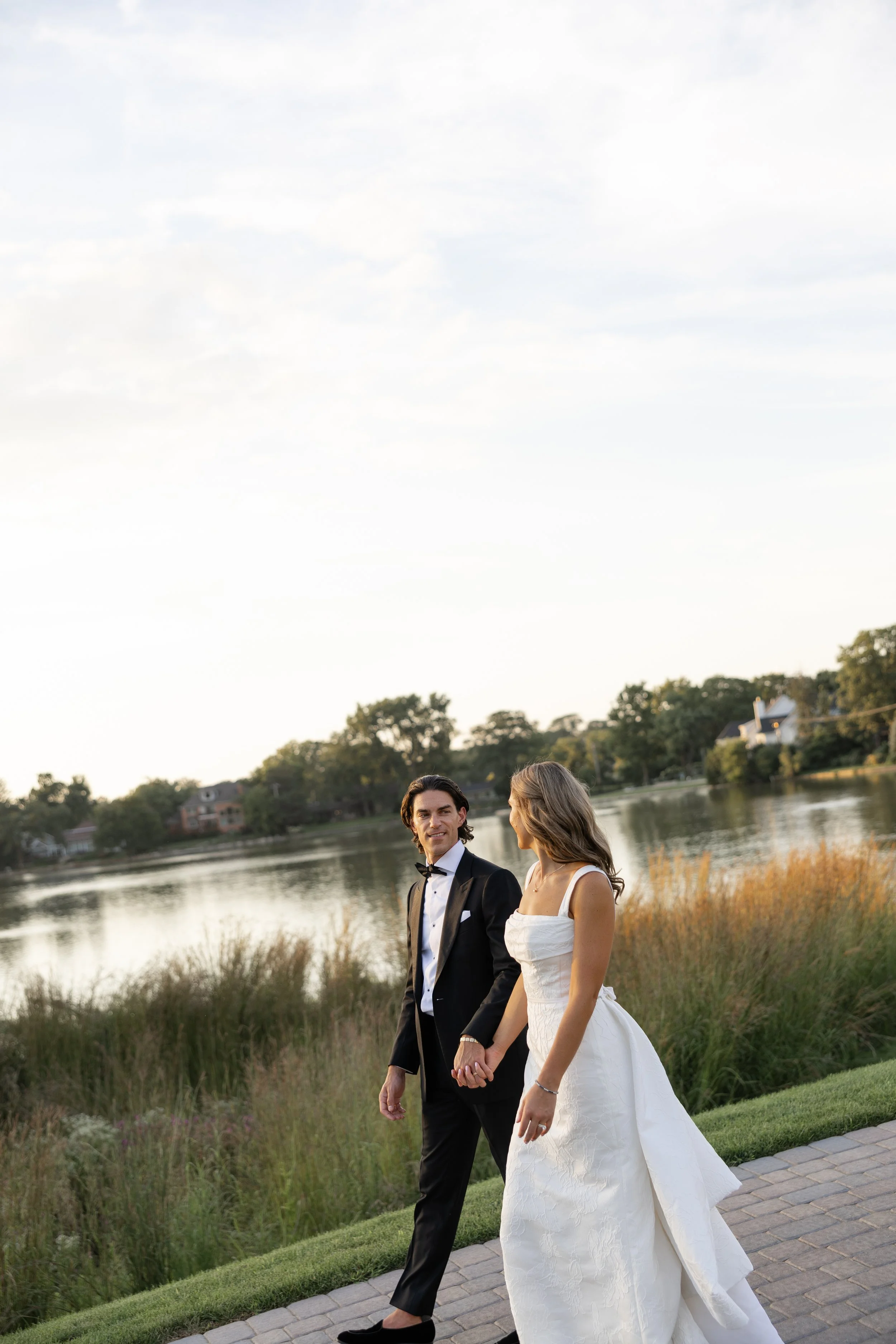A newlywed couple walking hand in hand on a pathway by a lake, with trees and houses in the background during sunset.