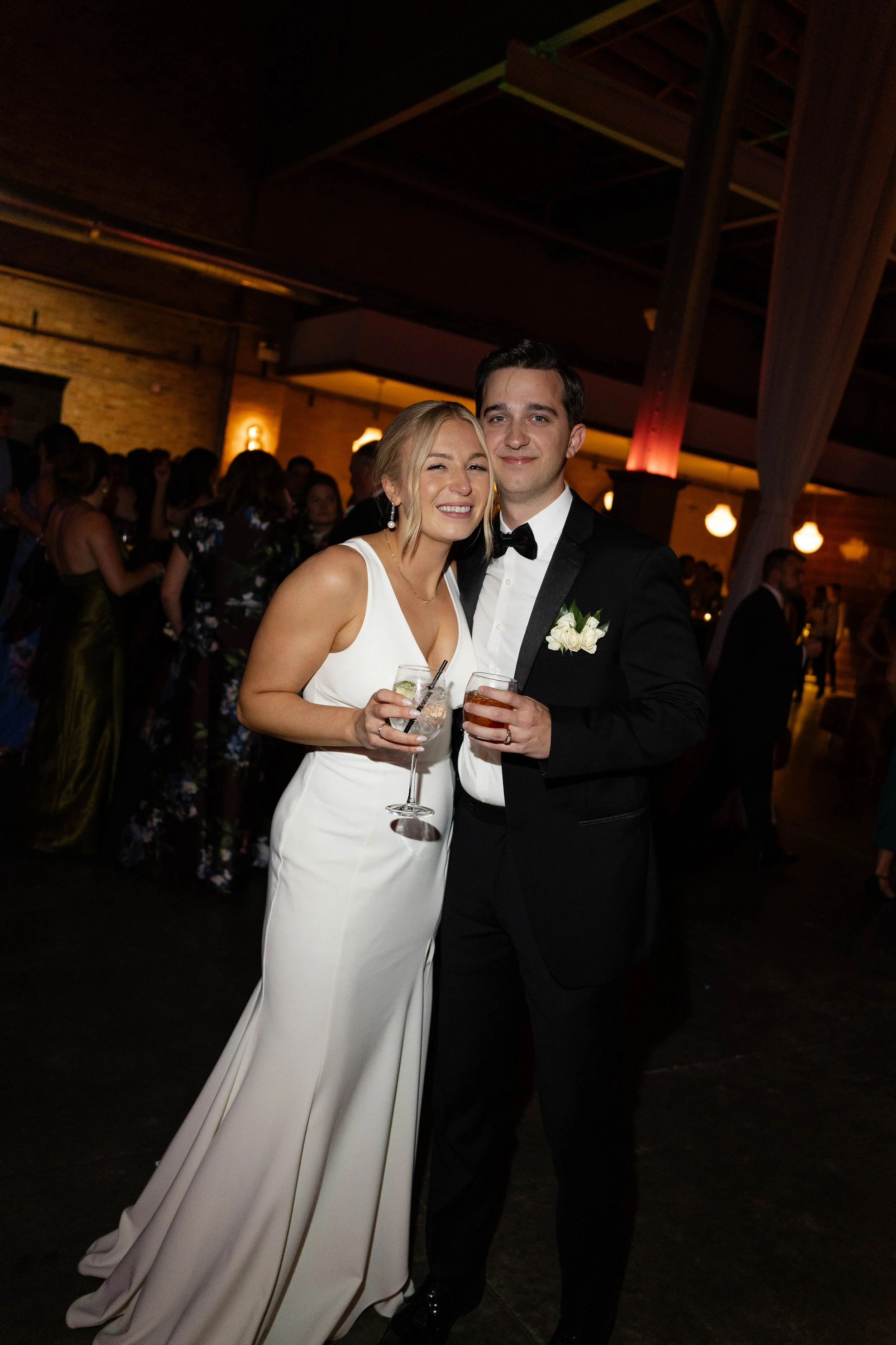 A smiling woman in a white dress and a man in a black tuxedo holding drinks at a formal event in a dimly lit venue with other guests in the background.