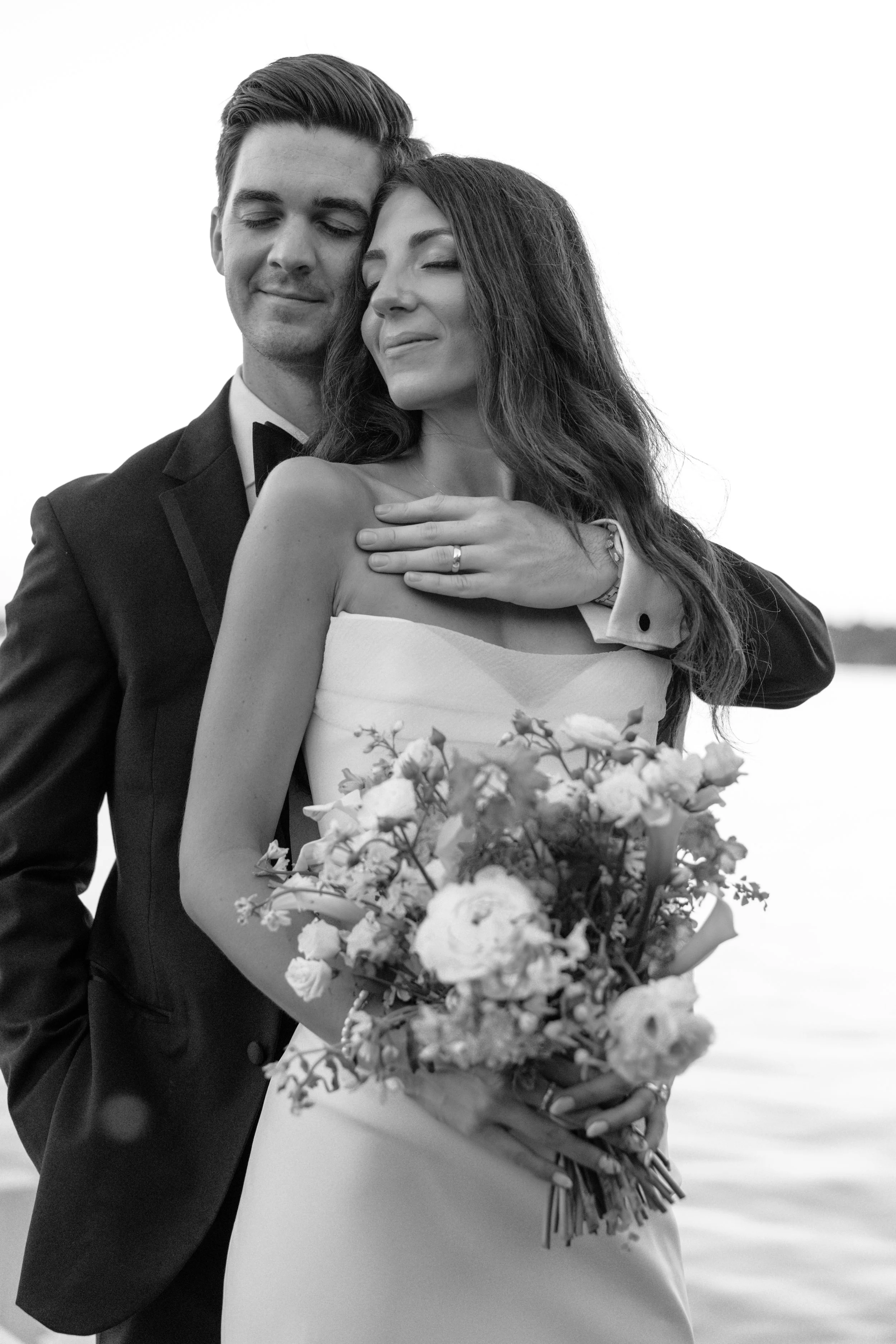 A black and white photo of a bride and groom embracing, with the groom in a tuxedo and the bride wearing a white dress holding a bouquet of flowers.