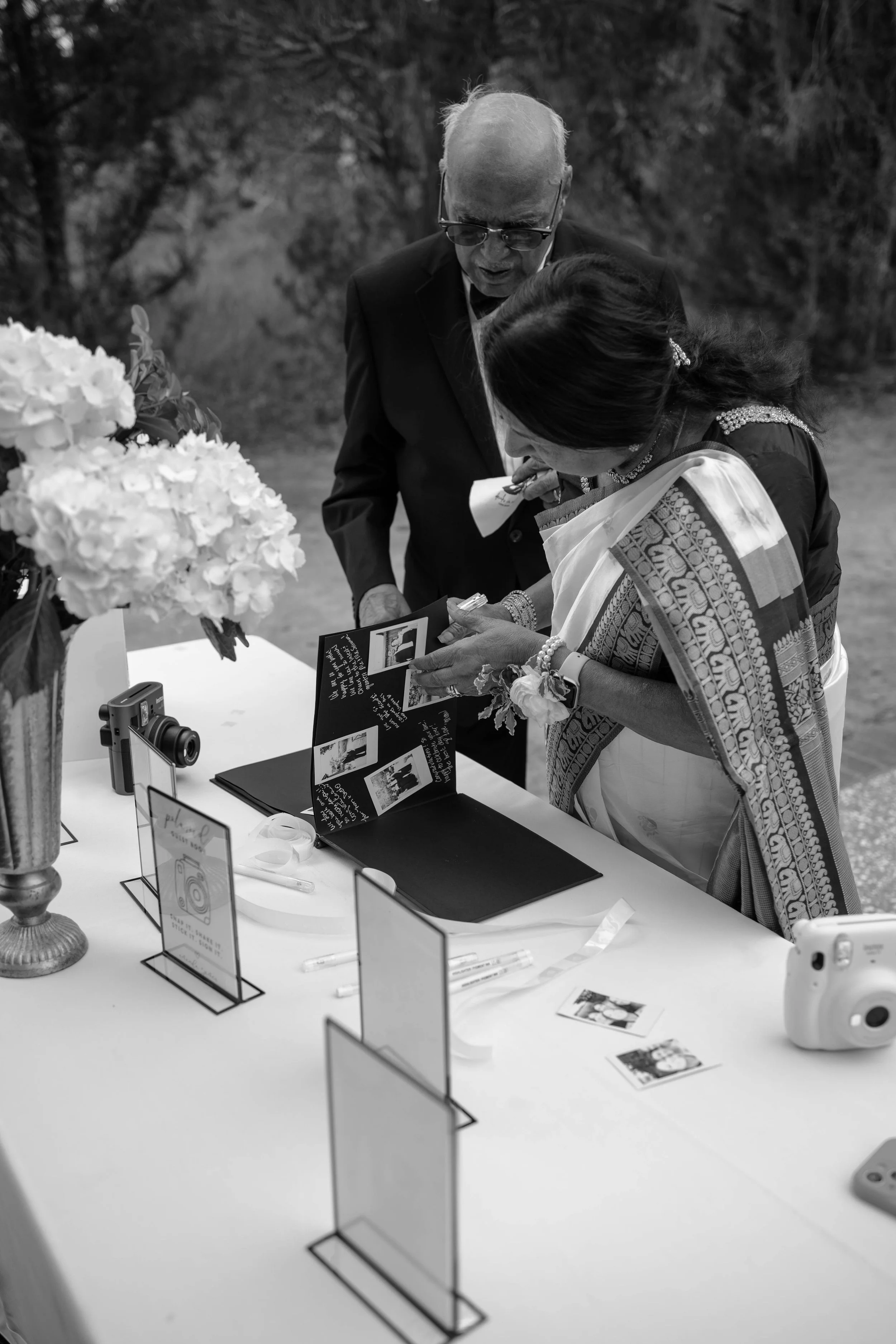 Two people with dark hair and glasses, an older man and a woman, looking at photographs and a booklet on a table decorated with flowers. There are small signs and a camera on the table.