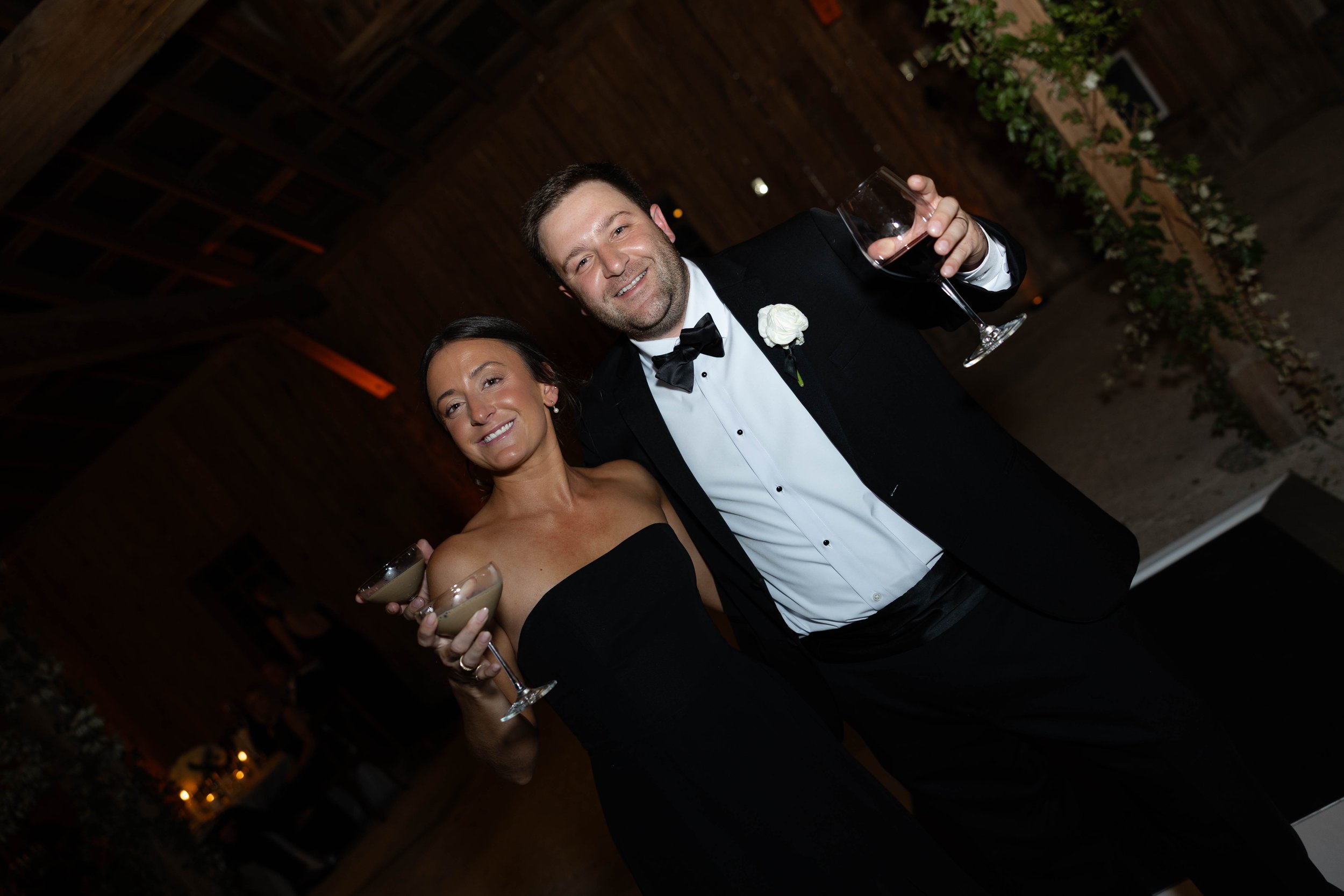 A man and woman dressed in formal attire, holding glasses of wine and smiling at an indoor event with wooden ceiling and greenery in the background.
