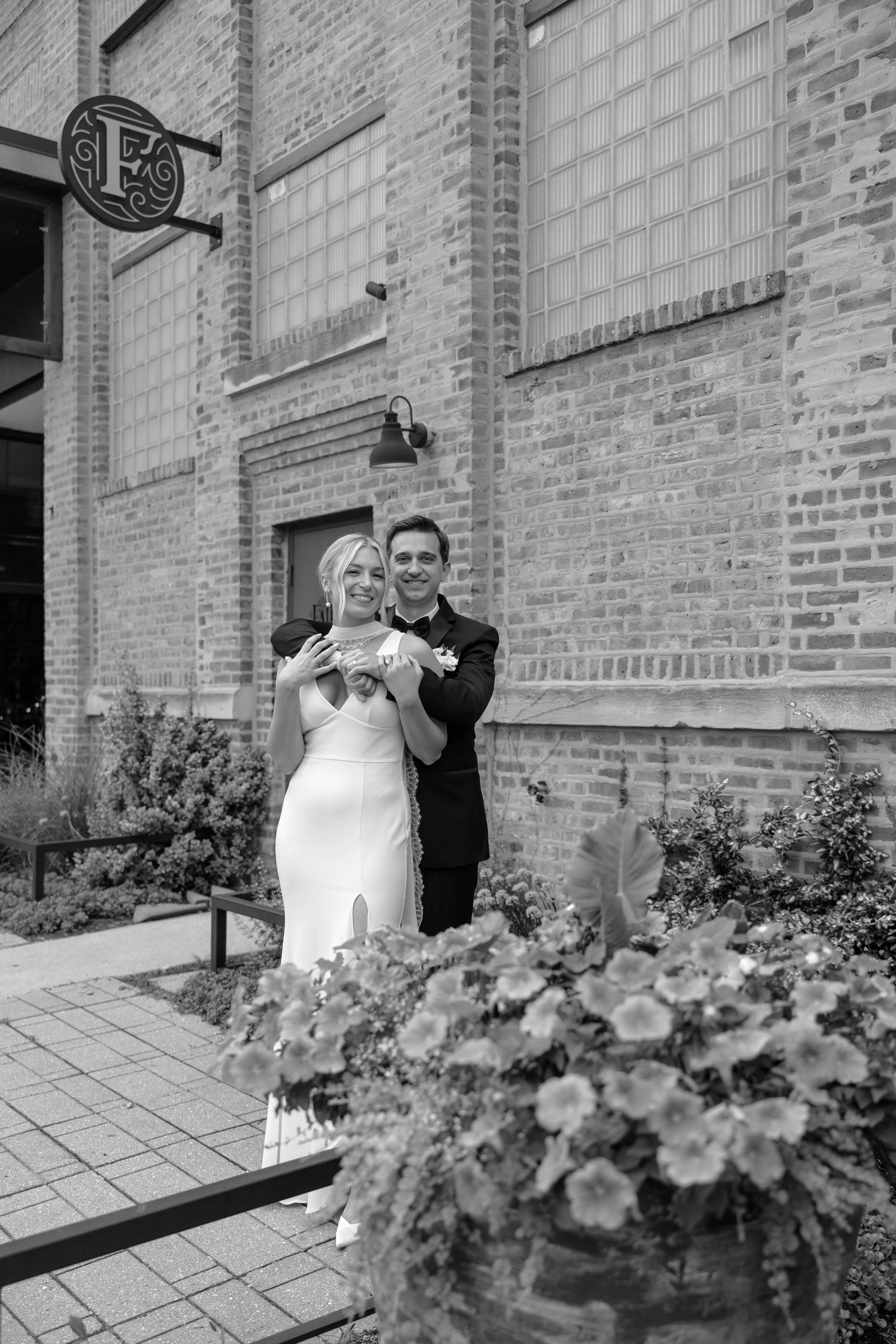 A bride and groom smiling and hugging outside in front of a brick building, with a flower bed in the foreground.