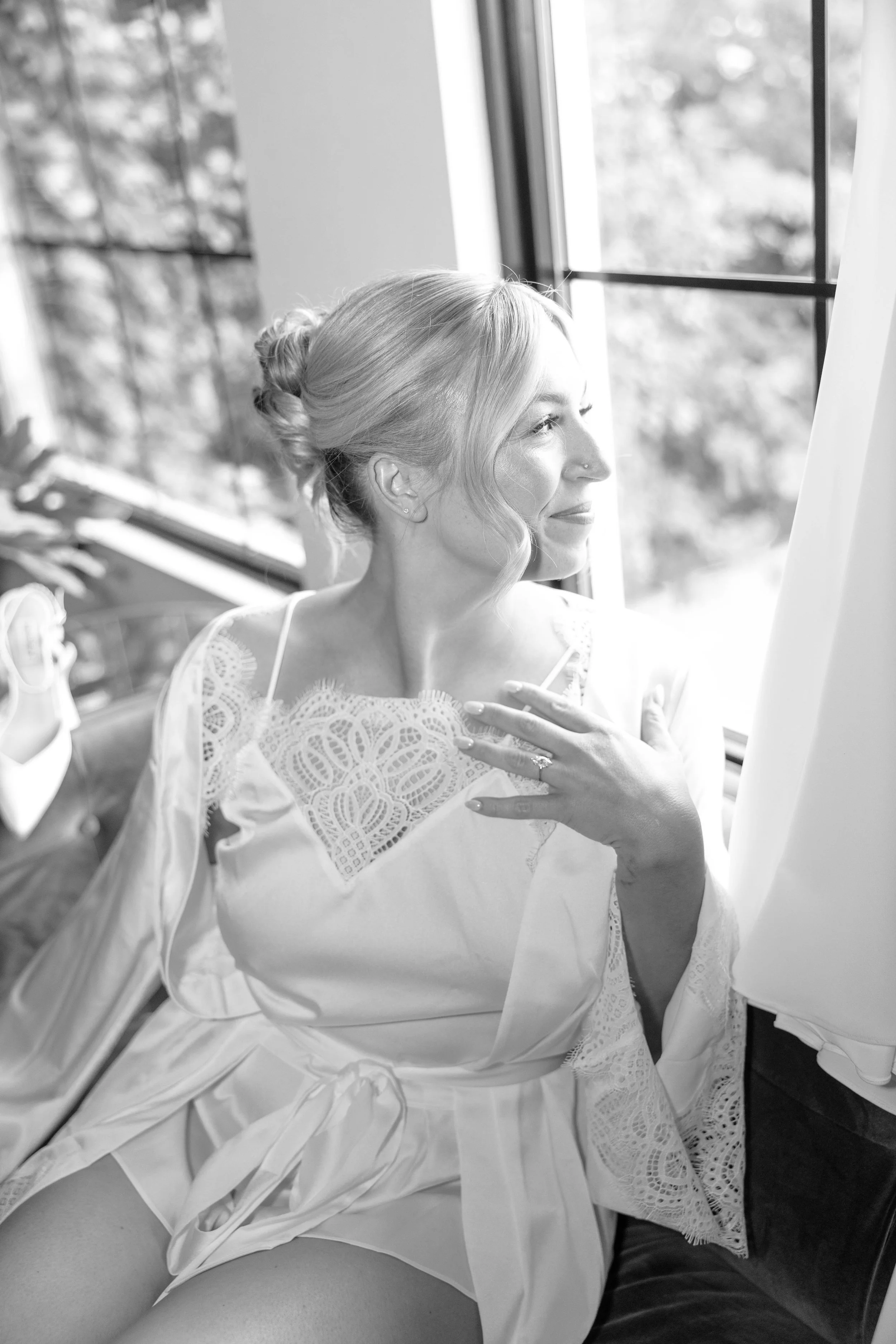 A woman with light hair in an elegant updo, wearing a lace dress, smiling and looking out a window in a black-and-white photo.