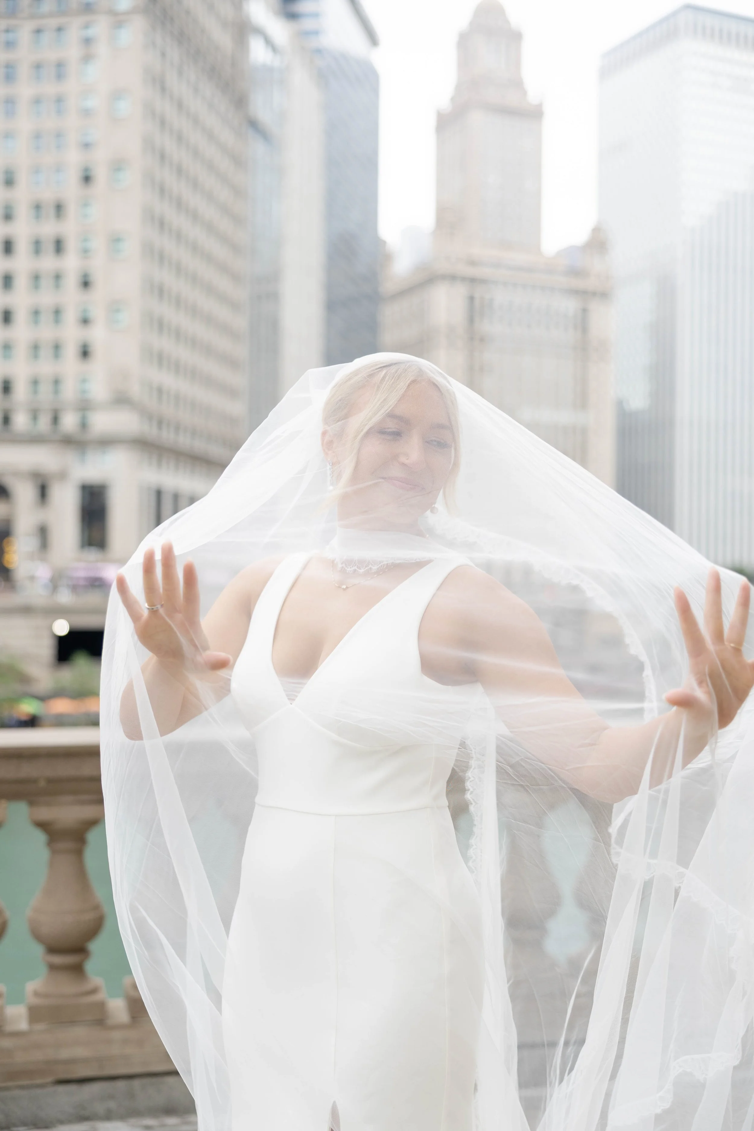 A bride in a white dress with a veil over her face, smiling and holding the veil up with both hands, standing on a balcony in an urban setting with tall buildings in the background.