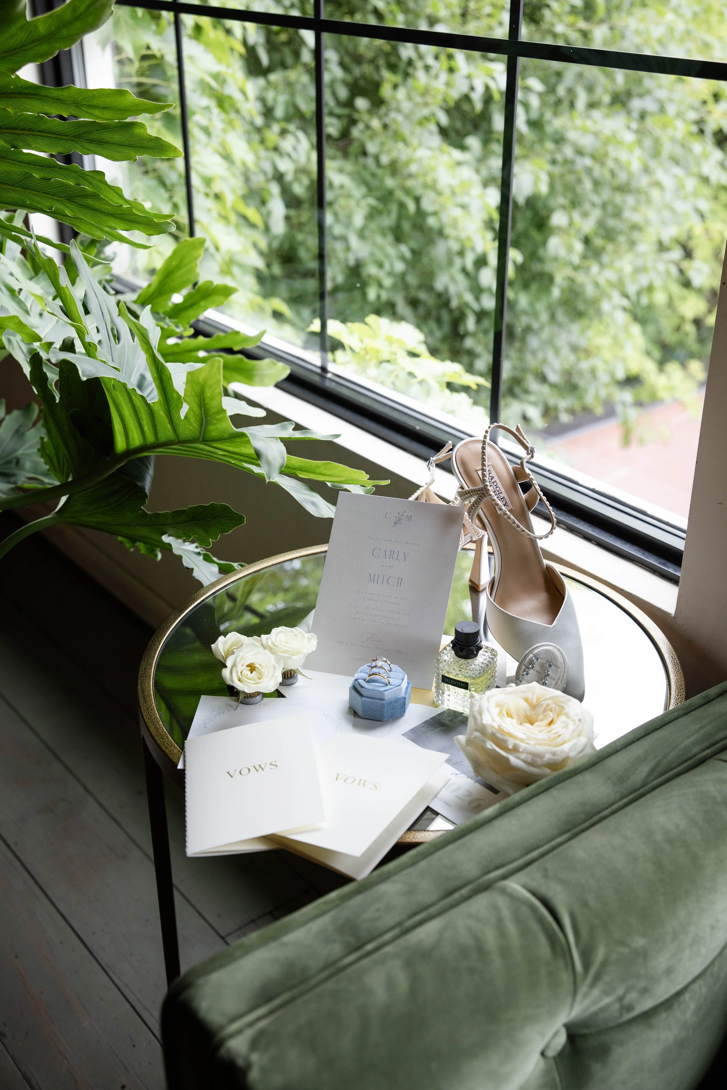 A small round table with white invitations, white roses, perfume, jewelry, and a pair of white high-heeled shoes next to a large green leafy plant and a window showing green trees outside.