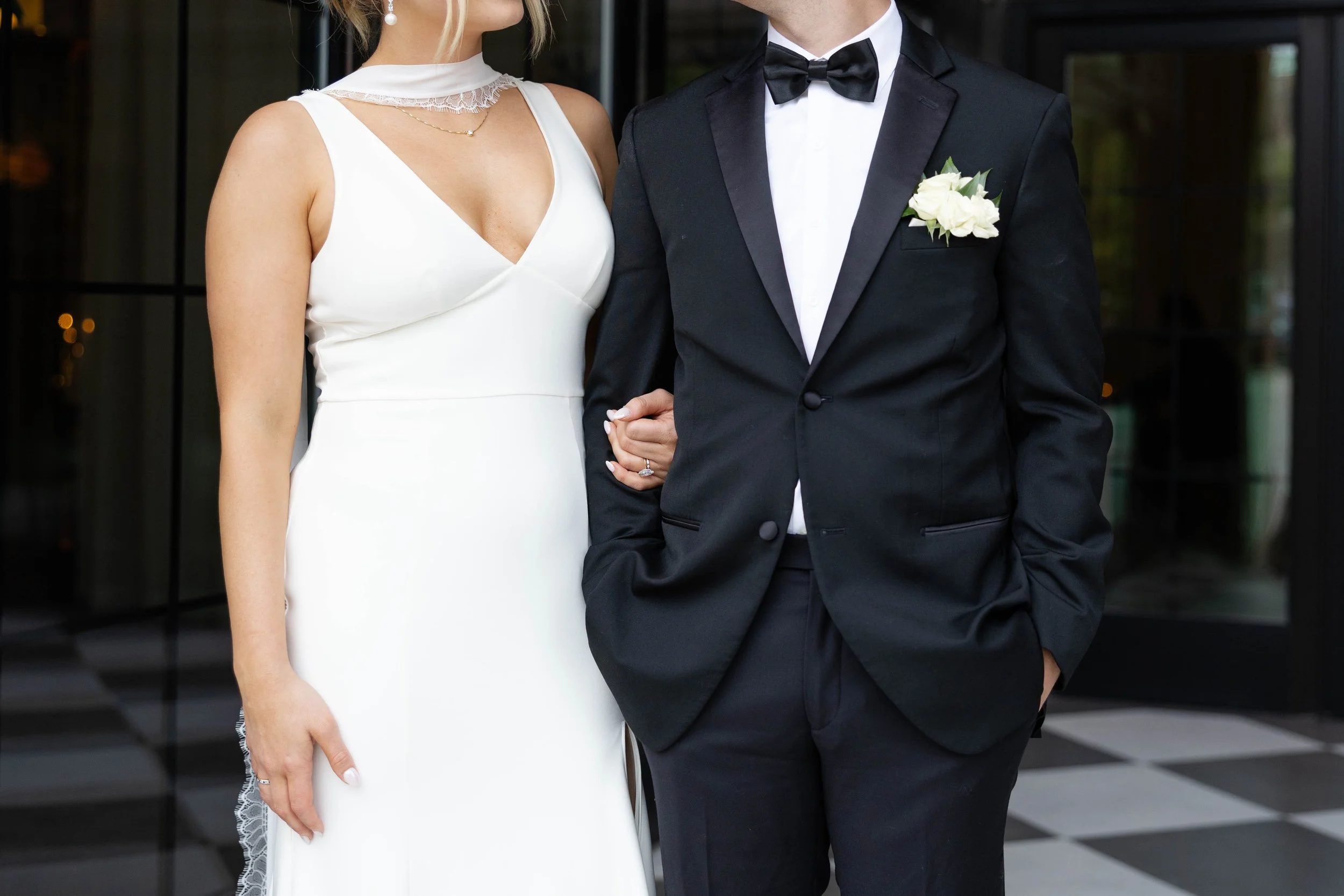 A bride and groom standing close together, holding hands. The bride is wearing a white wedding dress with lace details on the shoulders and a lace choker. The groom is dressed in a black tuxedo with a white shirt, black bow tie, and a white flower bo