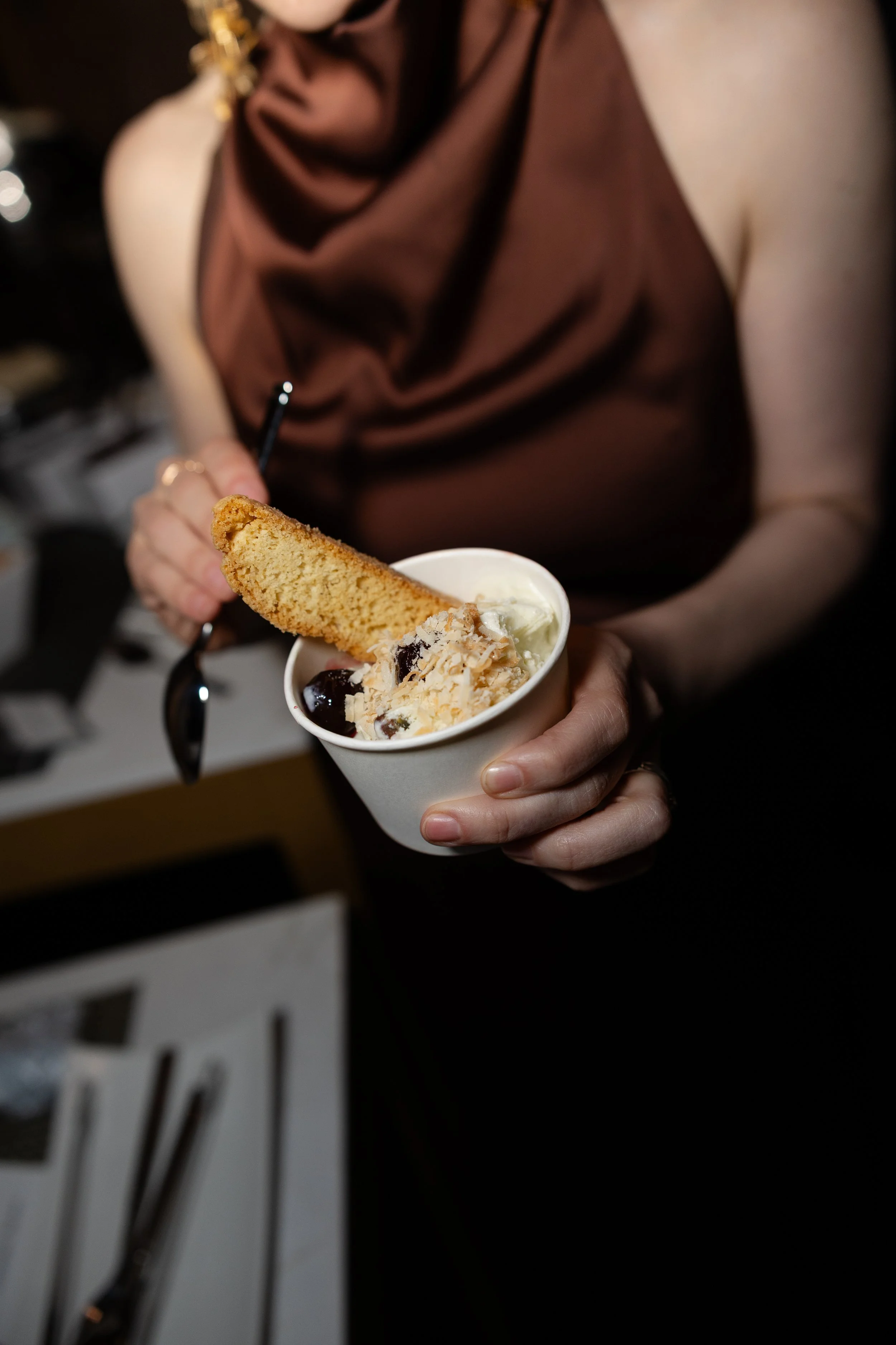 A woman in a brown sleeveless dress is holding a cup of ice cream with a cookie and some toppings, and is about to eat it.