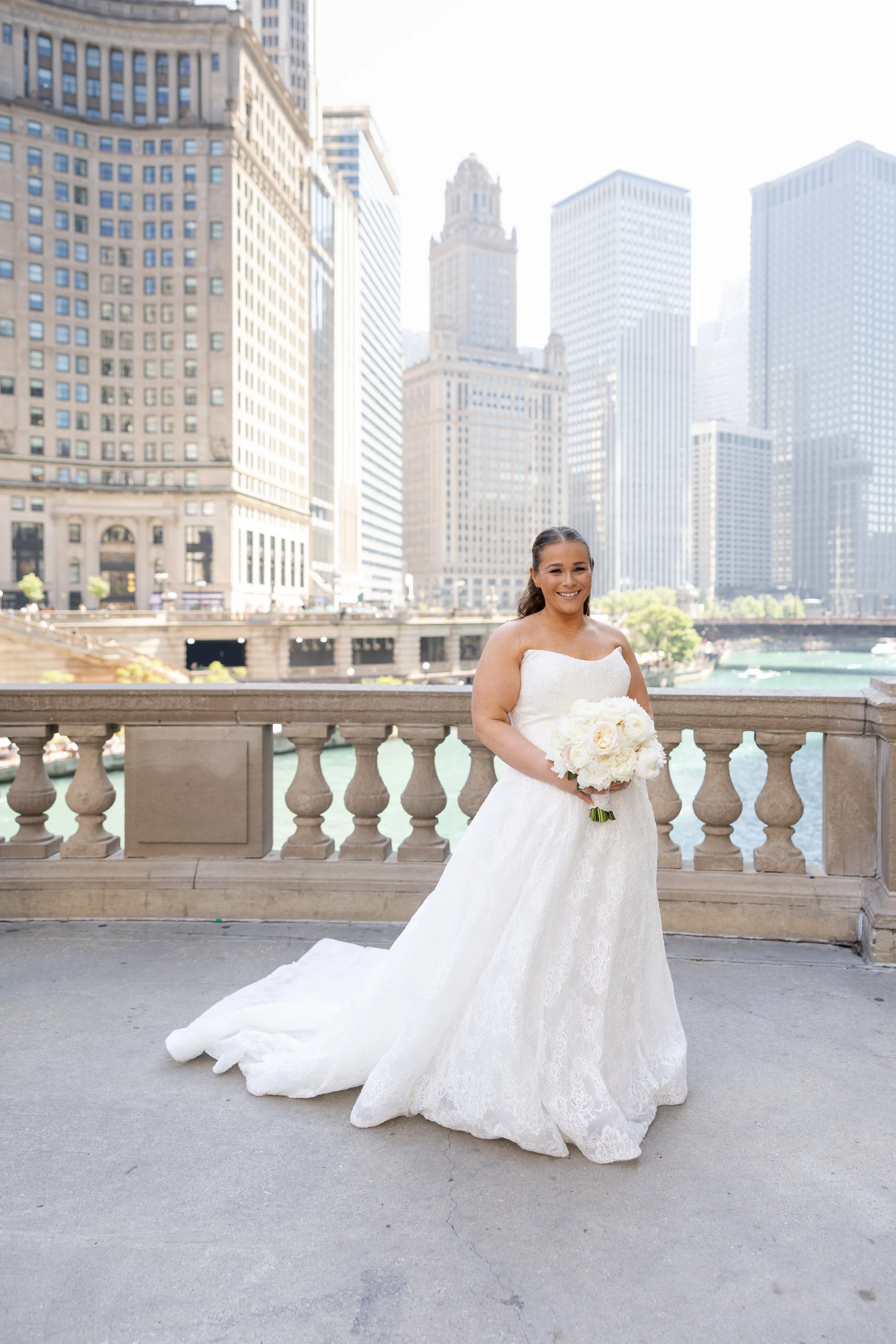 A woman in a white wedding dress holding a bouquet standing outdoors on a city bridge with tall buildings in the background.