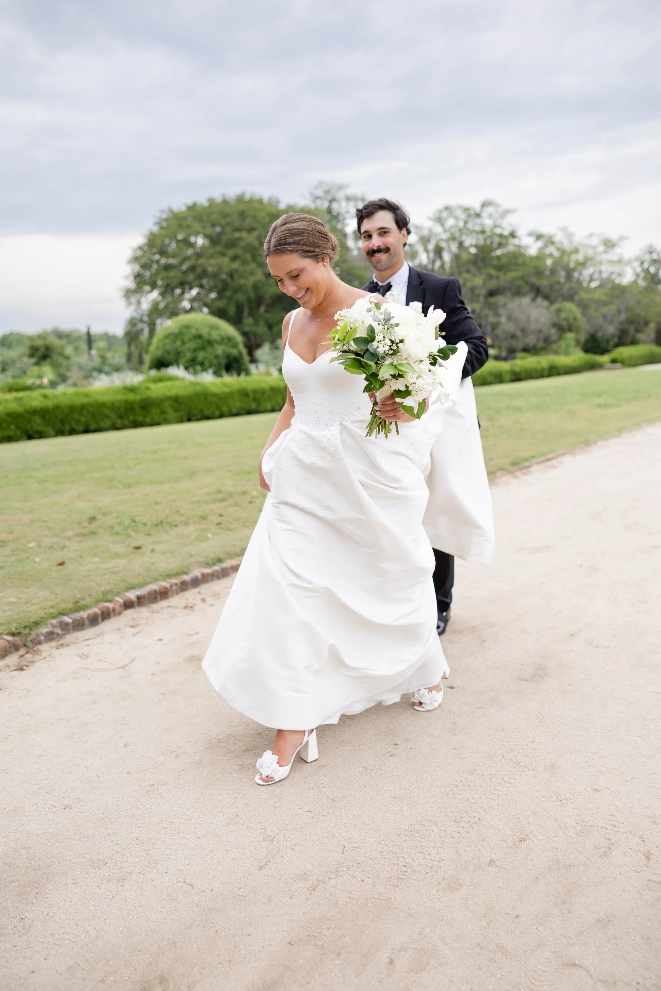 A bride in a white wedding dress holding a bouquet of white flowers and greenery, smiling as she walks on a dirt path with a groom following behind her on a cloudy day.