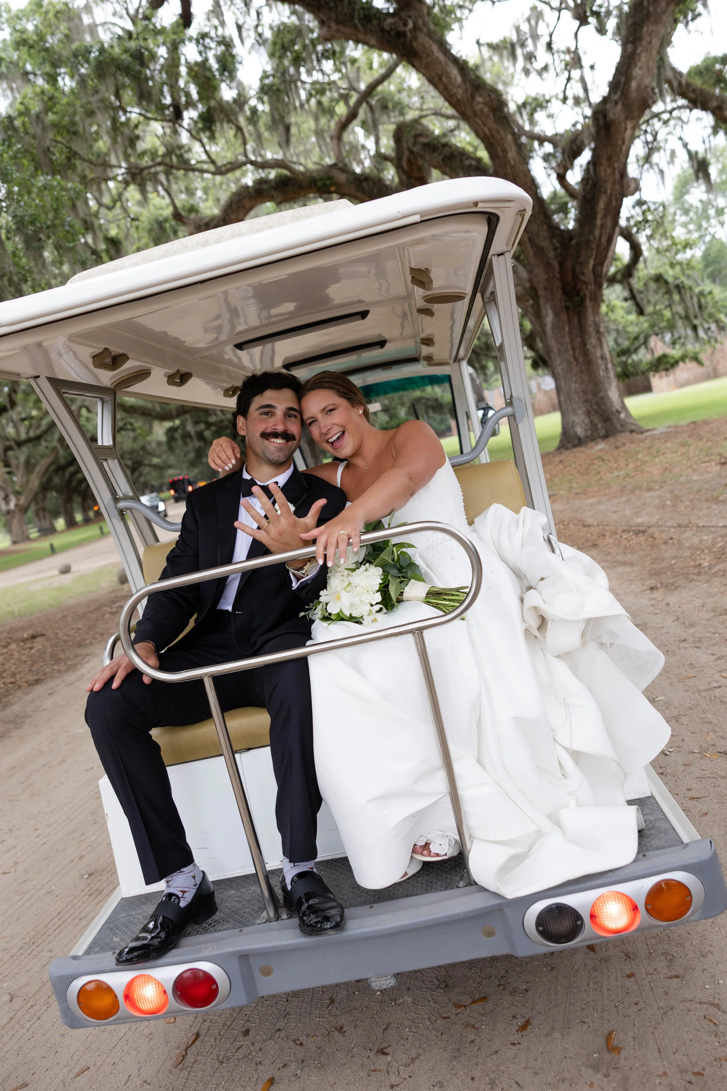 A bride and groom sitting in a golf cart, smiling and enjoying a joyful moment. The bride is wearing a white wedding dress and the groom is dressed in a black tuxedo. They are holding hands and appear happy.
