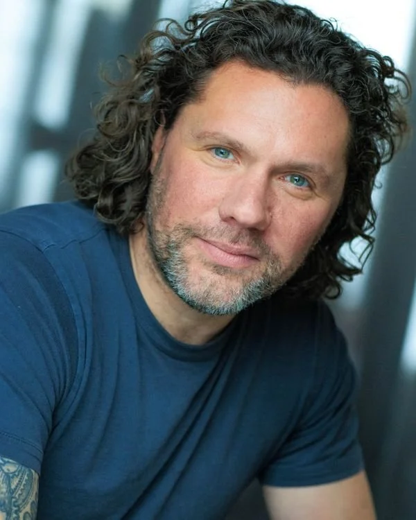 A man with curly dark hair and blue eyes, wearing a navy blue t-shirt, smiling softly at the camera.
