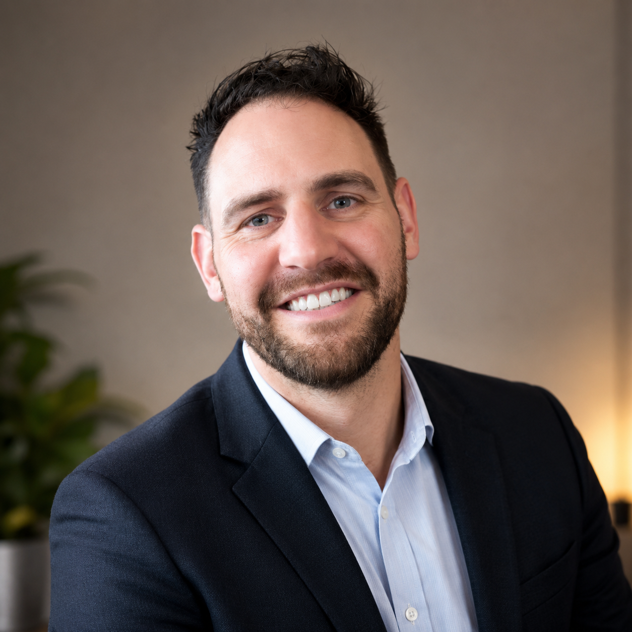 Professional headshot of a smiling man with dark hair and beard, wearing a navy blazer and light blue shirt, in an indoor setting with neutral background and a potted plant visible.
