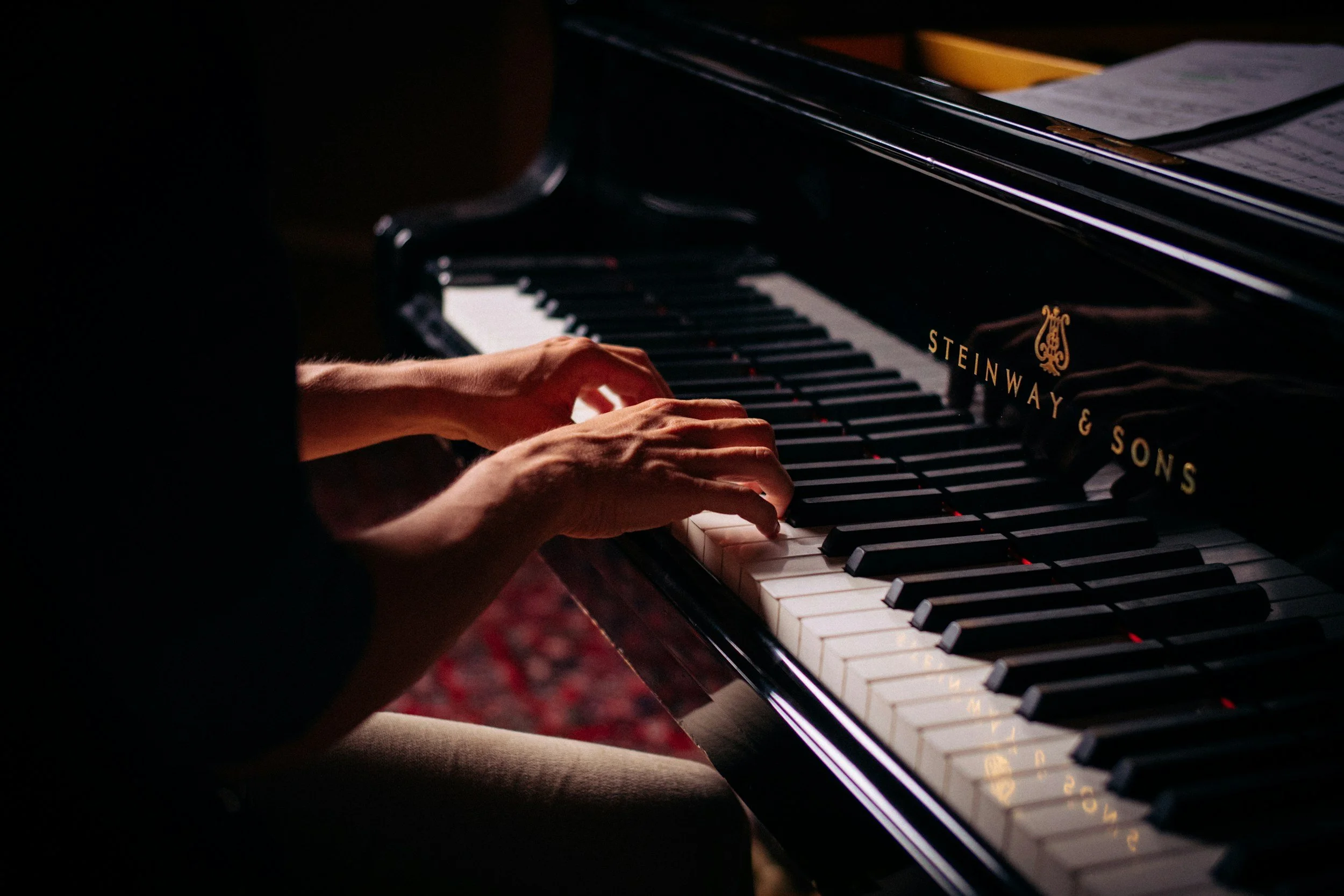 Close-up of hands playing a black Steinway & Sons grand piano in a dimly lit room.
