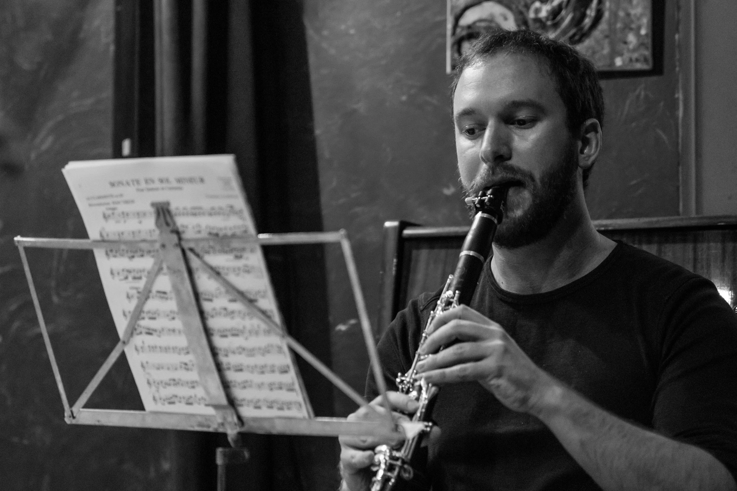 A man with a beard playing the clarinet and focusing on sheet music on a music stand in a dimly lit room with a dark wall and piece of abstract art hanging in the background.