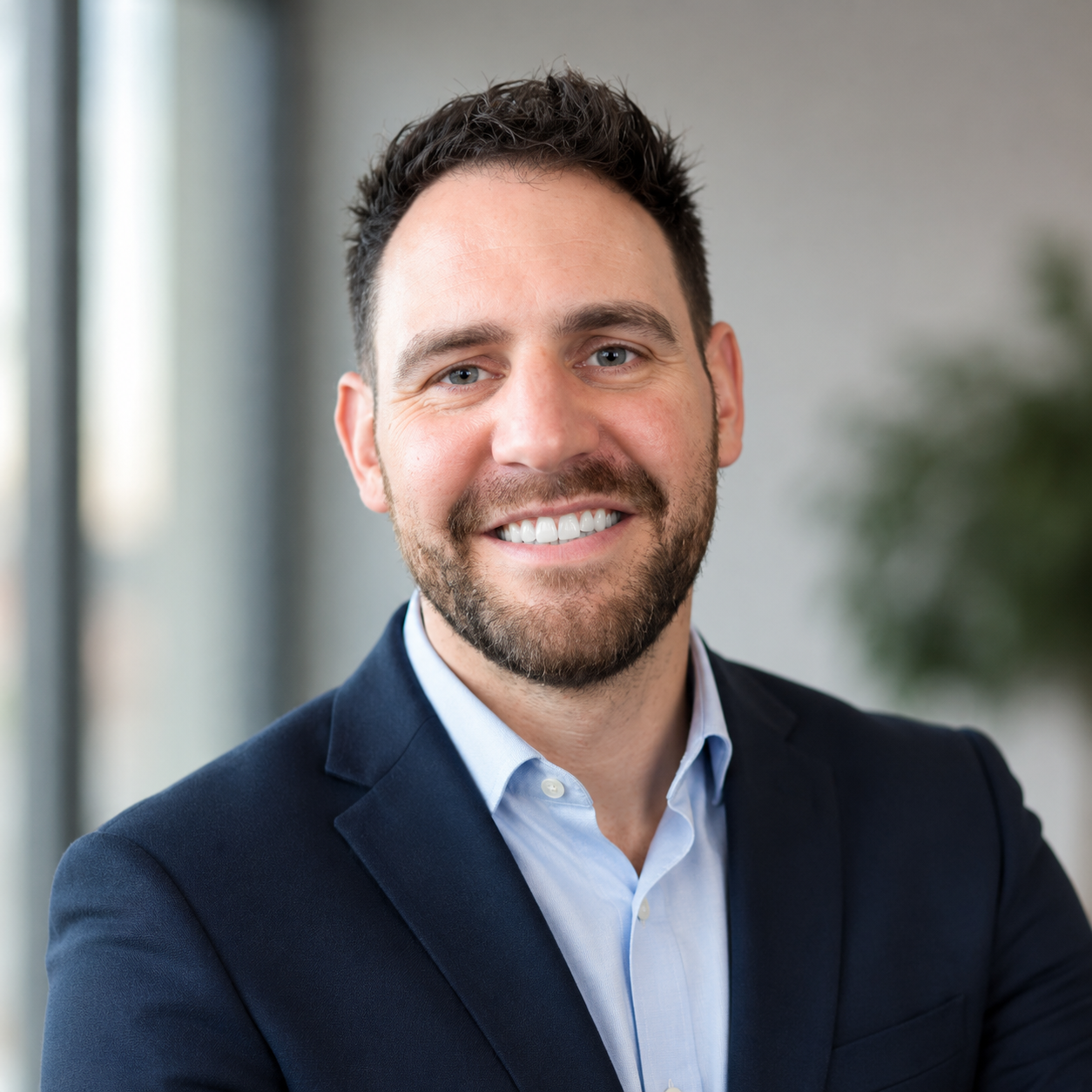 Headshot of a smiling man with dark hair and beard wearing a navy blazer and light blue shirt, in an office setting.