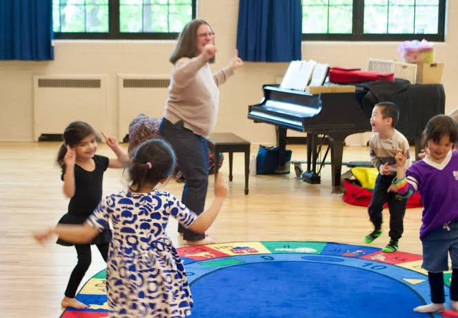 A teacher and four children dancing and having fun in a classroom with a blue circular rug.