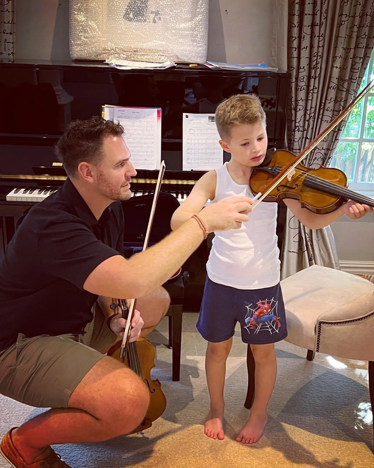 A young boy with a Spider-Man shirt and shorts playing the violin while an adult man kneels next to him, helping him hold the violin and bow in a room with a piano and musical sheets.