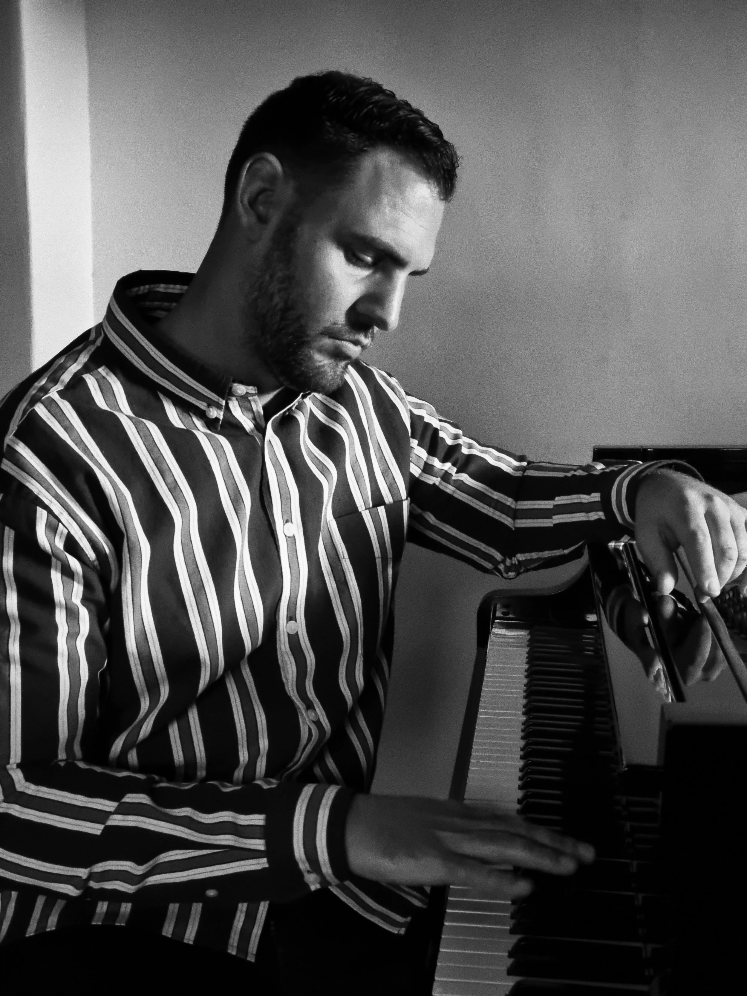 A black and white photo of a man playing a grand piano, wearing a striped shirt, with short dark hair and a beard, focused on playing the keys.
