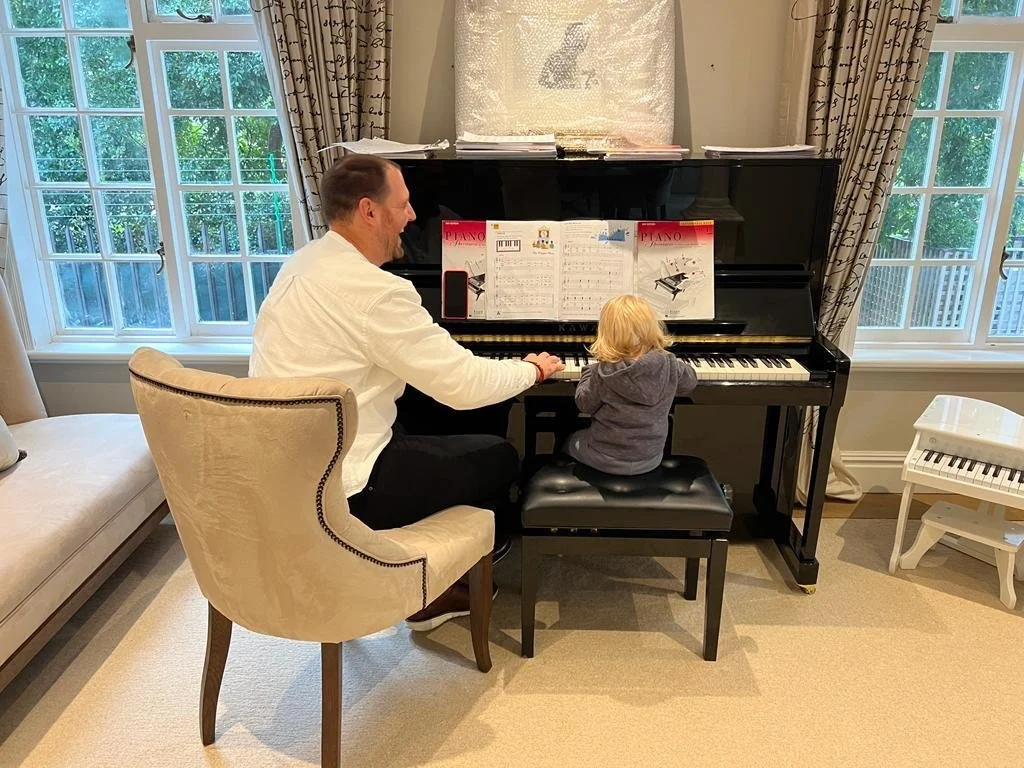 A man and a young child sitting at a black upright piano, with sheet music on the piano. The man is sitting in a beige chair and is holding the child's hand as they play the piano. The room has large windows with curtains on either side, and a beige carpet.