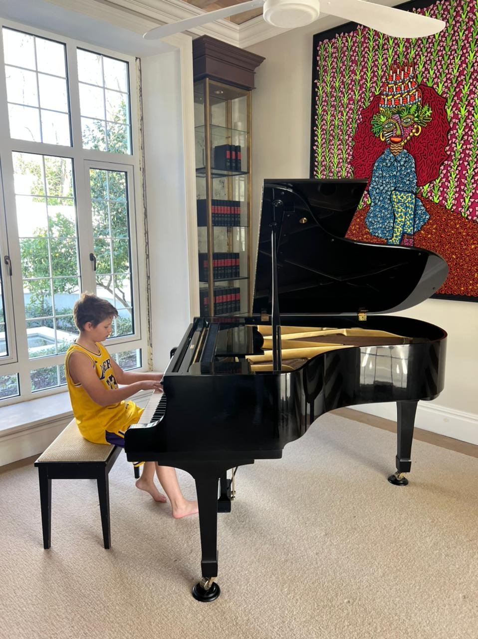 A young boy in a yellow basketball jersey sitting at a black grand piano in a room with large windows, a colorful artwork on the wall, and a glass display cabinet.