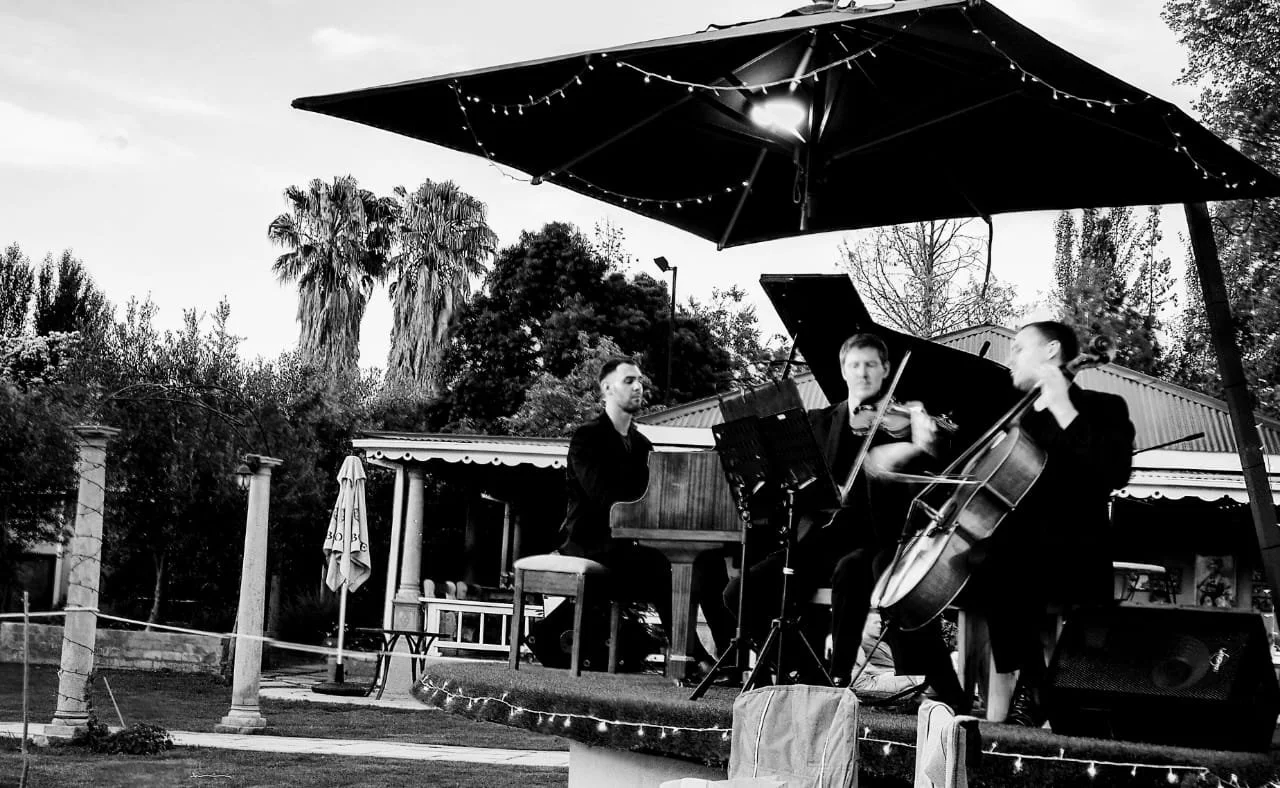 Three musicians performing outdoors under a large umbrella with string lights, including a pianist, a violinist, and a cellist.