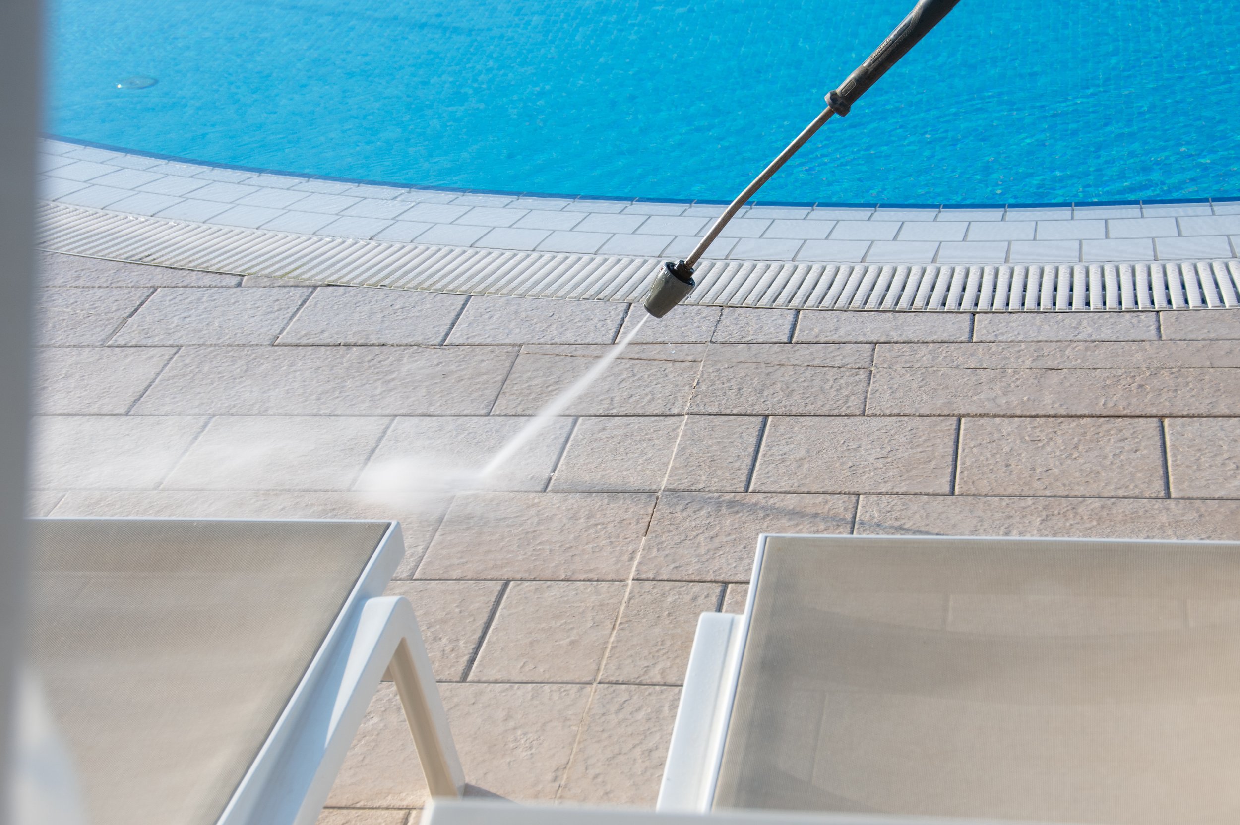 Poolside scene with a vacuum cleaner on beige stone tiles near a blue swimming pool. Two lounge chairs are partially visible in the foreground.