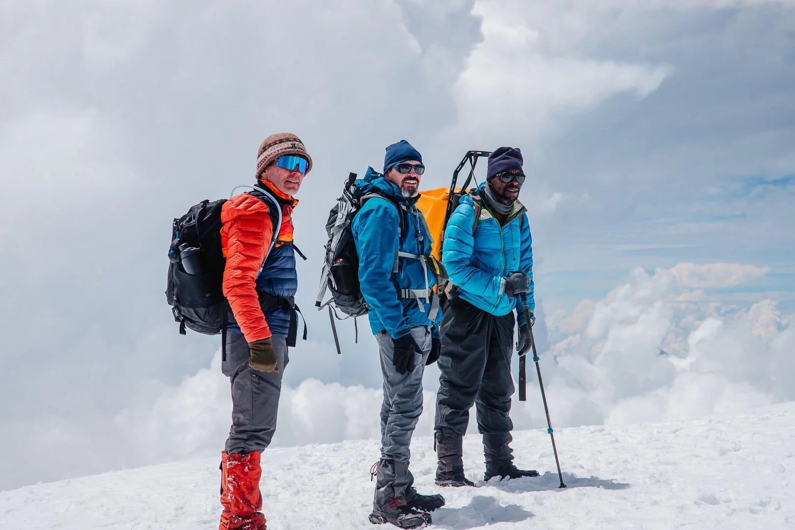 Three people dressed in winter climbing gear standing on snowy mountain summit with cloudy sky in the background.