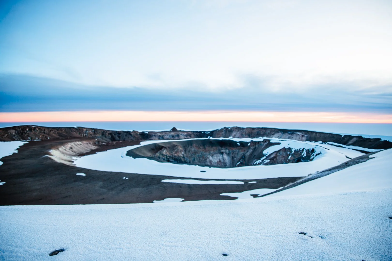 A volcanic crater filled with snow, surrounded by rugged hills and a partly cloudy sky at sunset or sunrise.