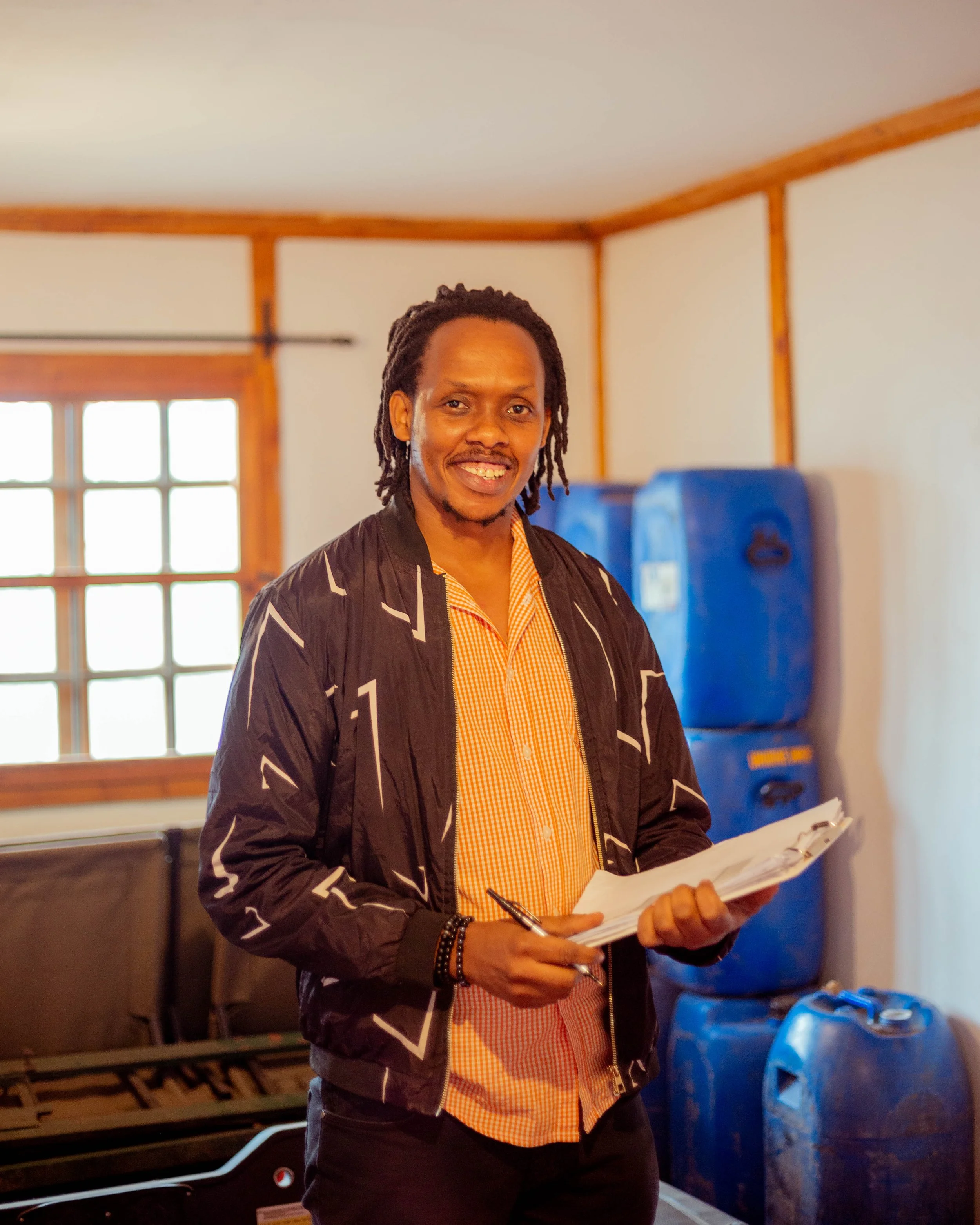 A man with dreadlocks smiling, holding a clipboard and pen, standing in a room with wooden trim, large window, and blue recycling bins in the background.