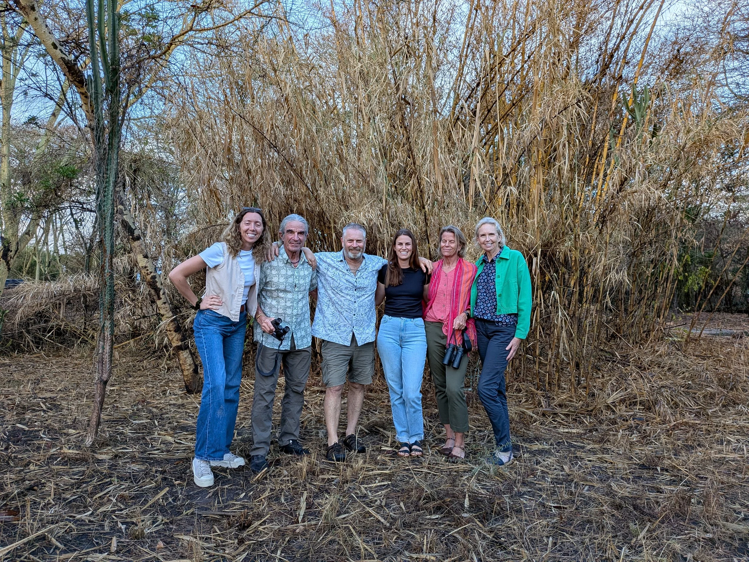 Group of seven people standing outdoors in front of dry, tall grass in a natural setting.