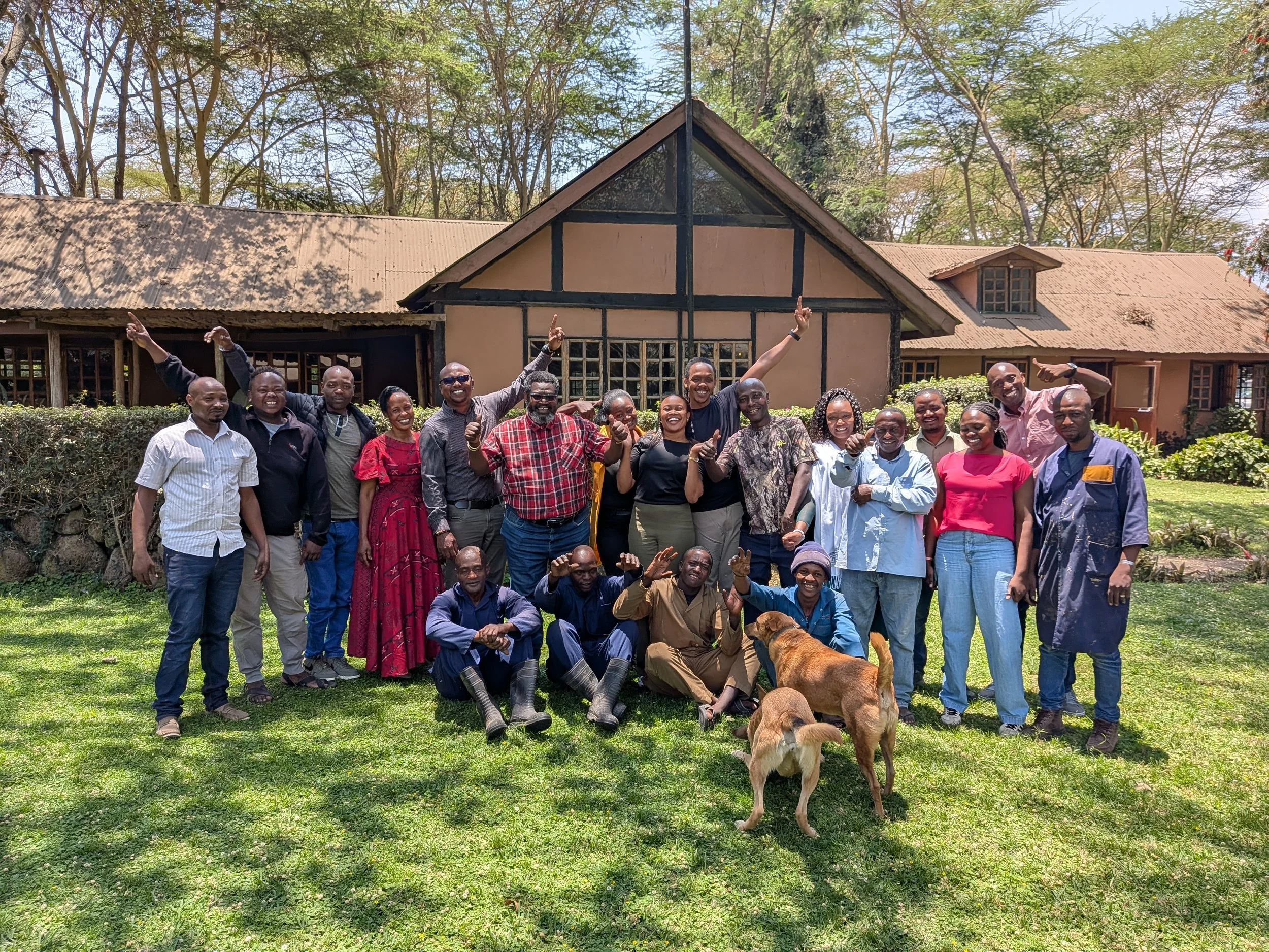A group of people standing outdoors on grass, smiling and posing for a photo, with a house and trees in the background. Dogs are lying on the ground in front of the group.