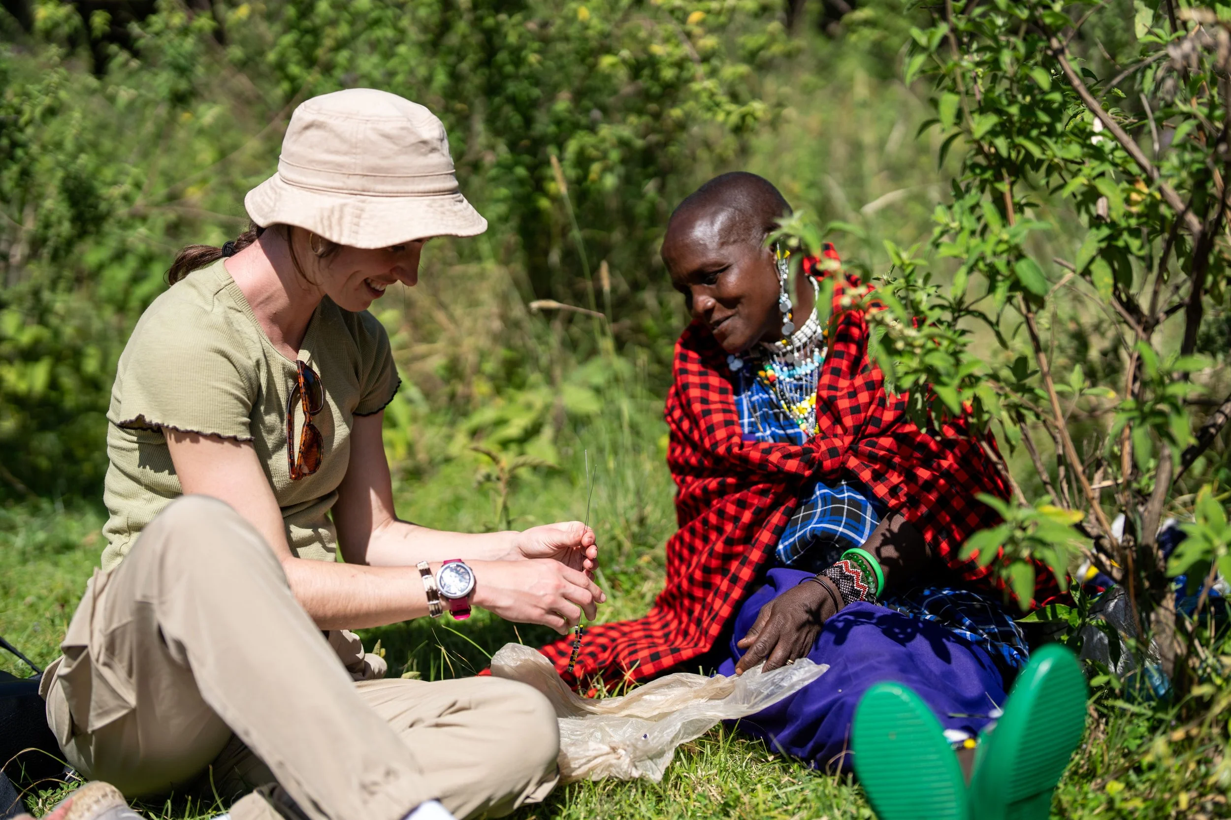 Two women, one in light clothing and a beige hat, and the other in traditional Maasai attire with jewelry, sit outdoors among green bushes, engaging in a conversation or activity, smiling and enjoying each other's company.