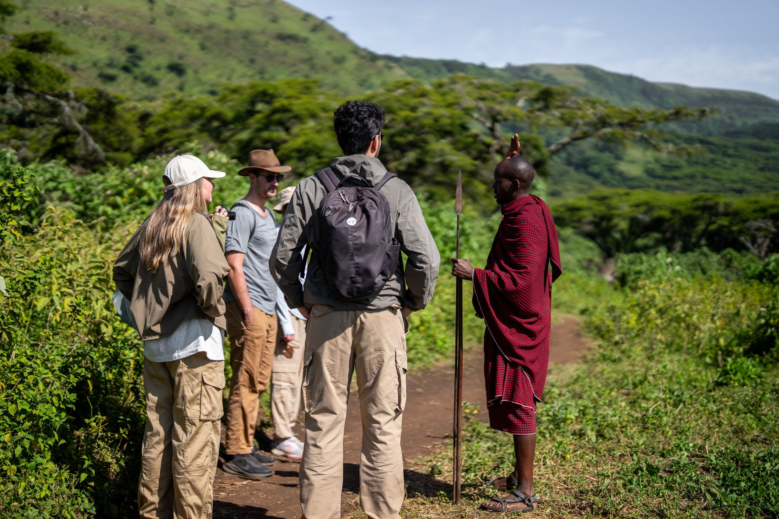 A group of tourists standing on a dirt path listening to a local guide dressed in traditional Maasai attire holding a spear in a lush, green landscape.