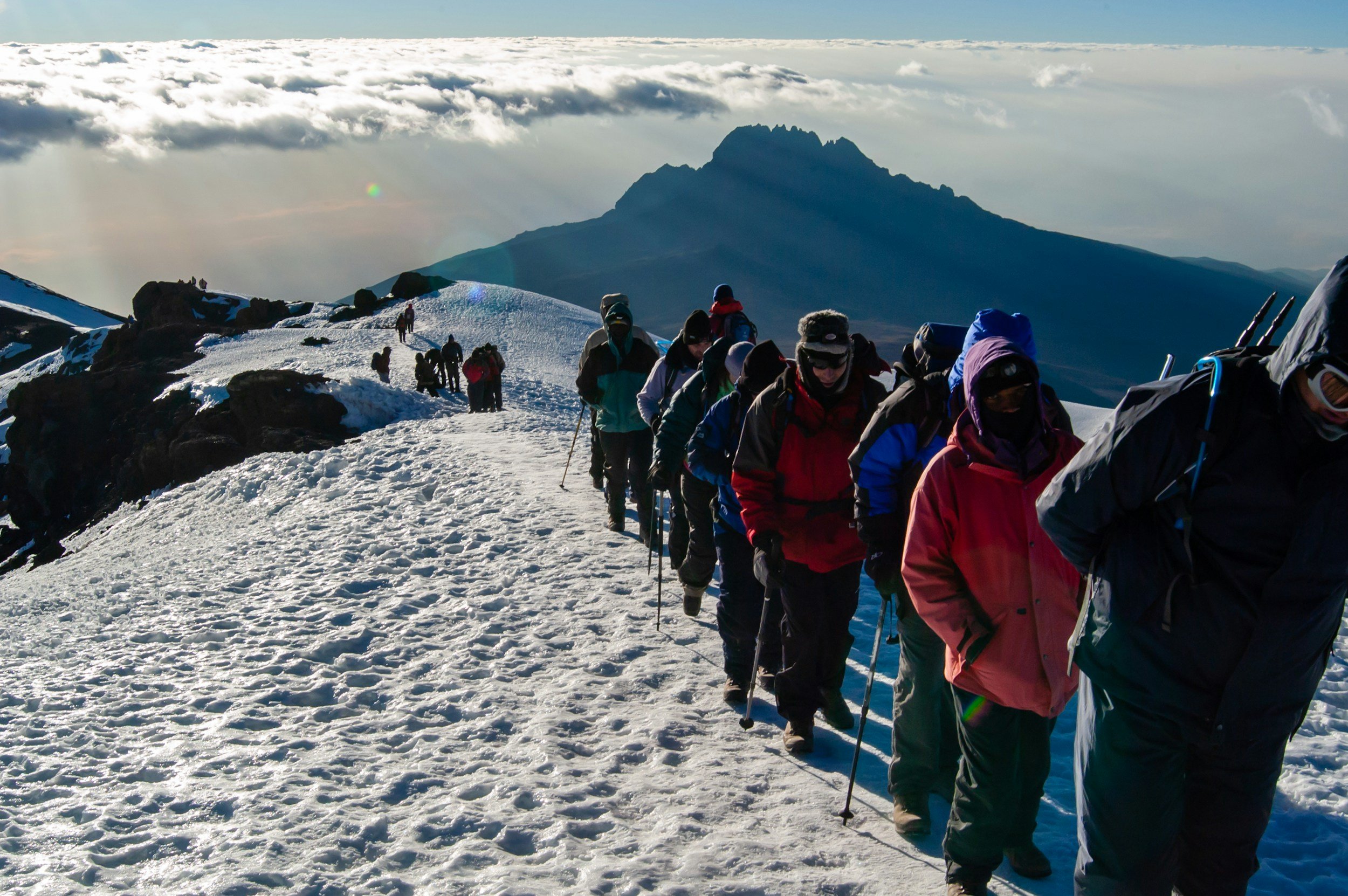 Group of people hiking on a snowy mountain trail with a mountain peak in the distance under a partly cloudy sky.