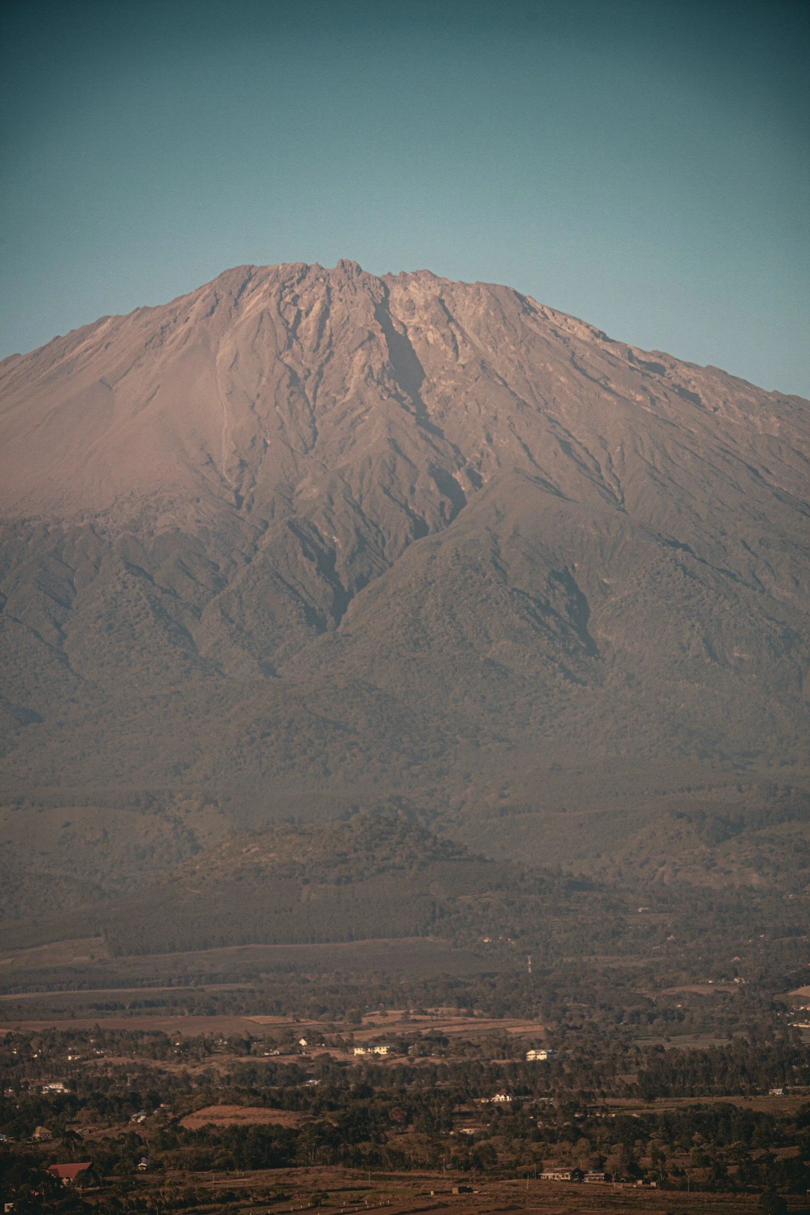 A large mountain with rugged slopes and a snowless peak stands in the distance, with a clear blue sky above. The foreground shows a flat landscape with patches of farmland, scattered houses, and trees.