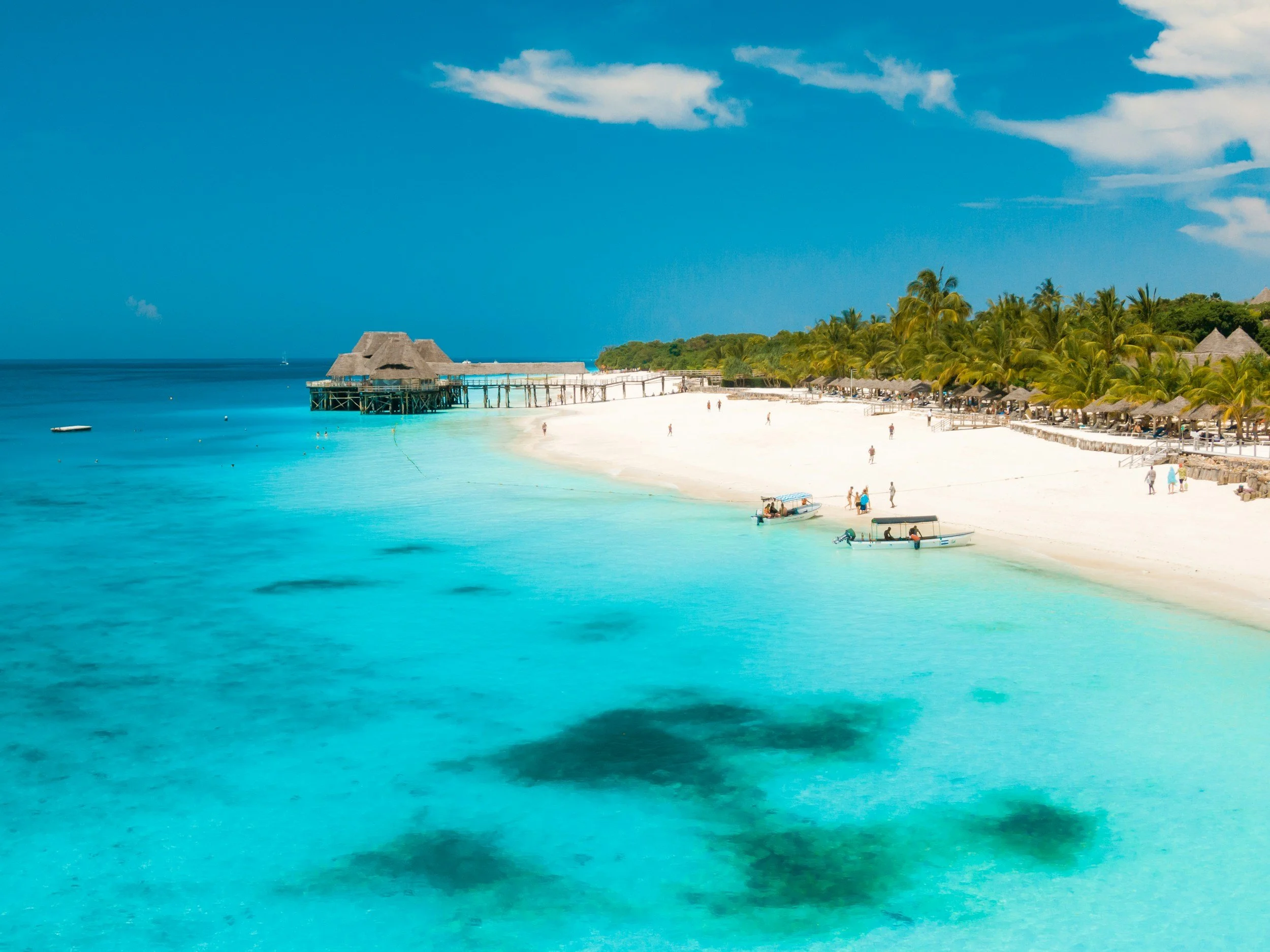 A tropical beach with turquoise water, white sand, palm trees, and thatched-roof huts along the shoreline. A wooden pier extends over the water with overwater bavilions.