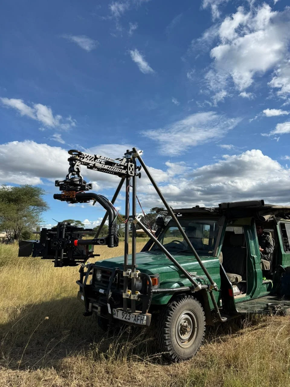 A green off-road vehicle equipped with a camera rig and mechanical arm for wildlife filming, parked on a grassy field under a partly cloudy sky.