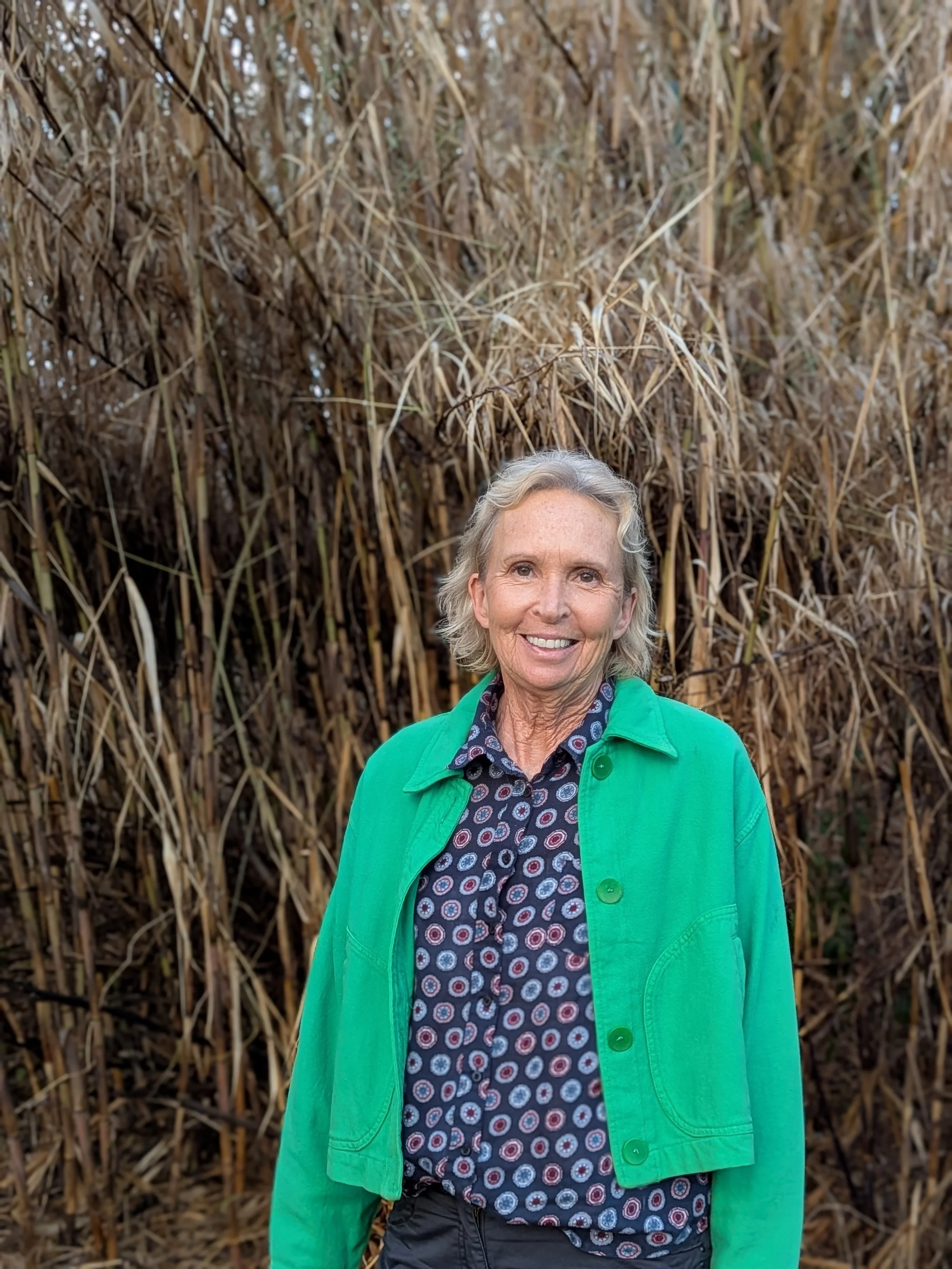A smiling woman with gray hair wearing a green jacket over a patterned shirt, standing in front of dry, tall grass or reeds.