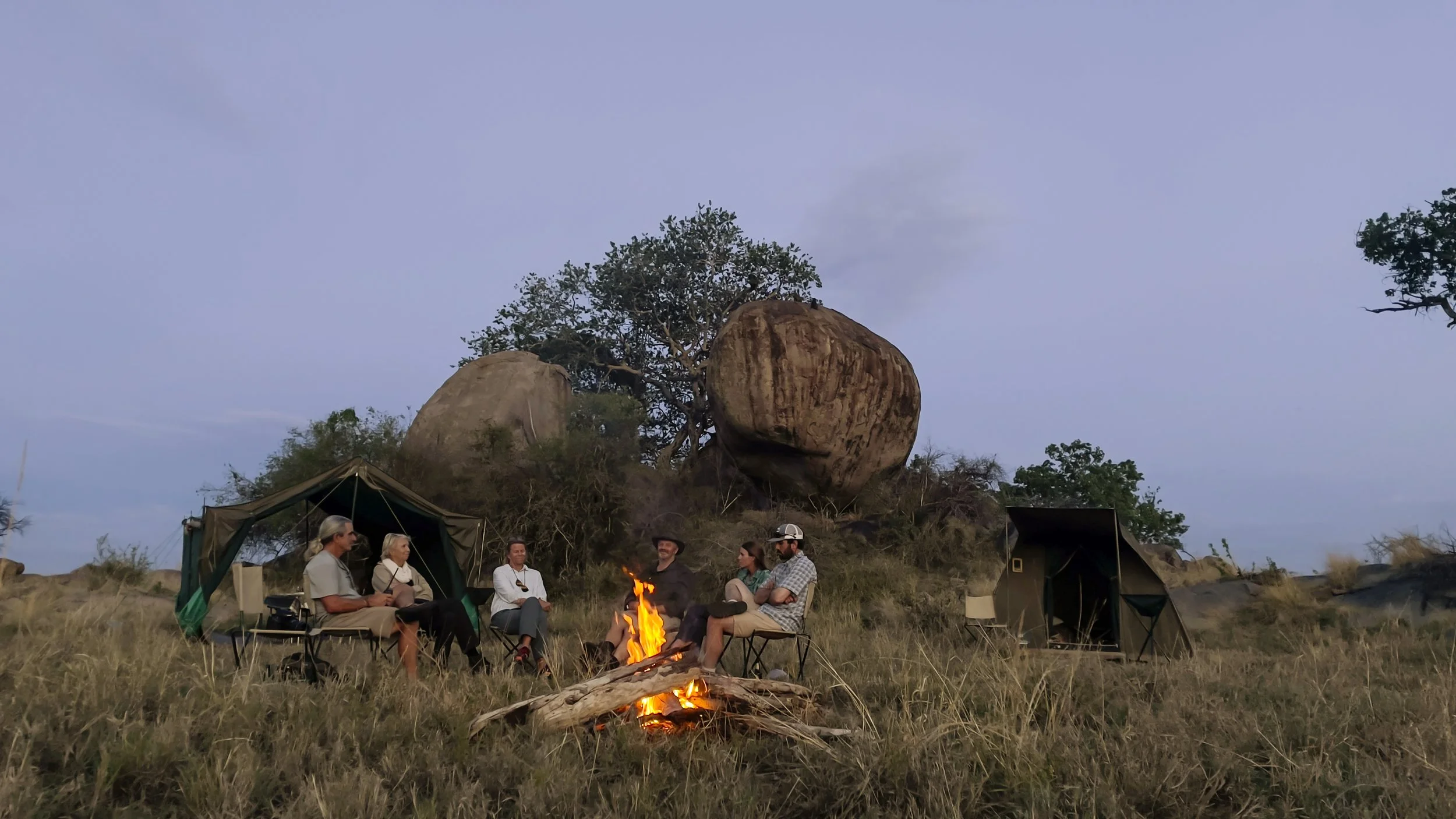 Group of people sitting around a campfire, camping tents in a grassy area with large rocks and trees, during dusk or early evening.